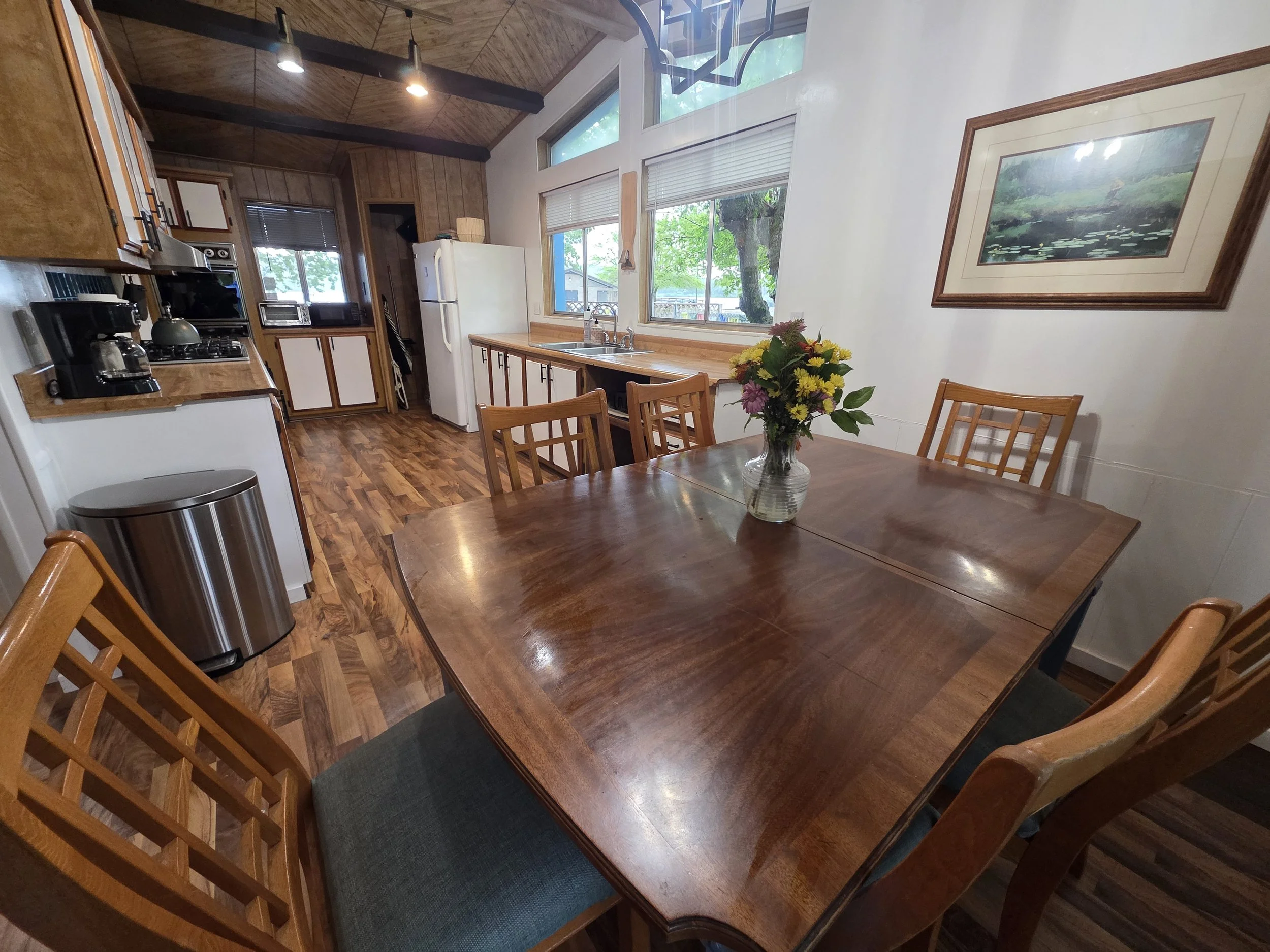 A cozy kitchen and dining area with a wooden table and six chairs, a vase of flowers, wooden cabinetry, and a window showing trees outside.