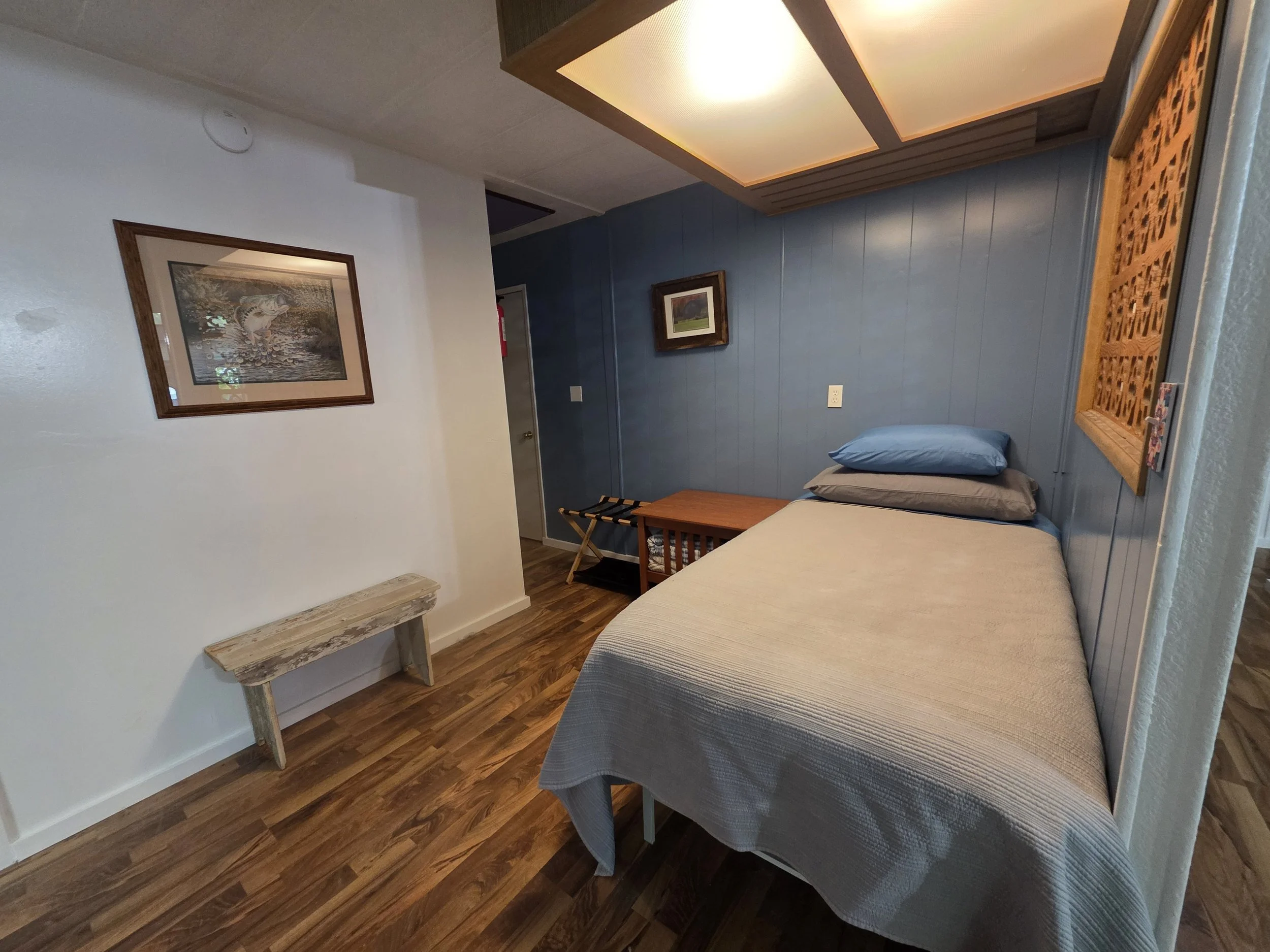 Room with a bed covered by a white bedspread, blue and gray pillows, wooden side table, framed pictures on the walls, wood paneling, and a patterned wooden wall panel.