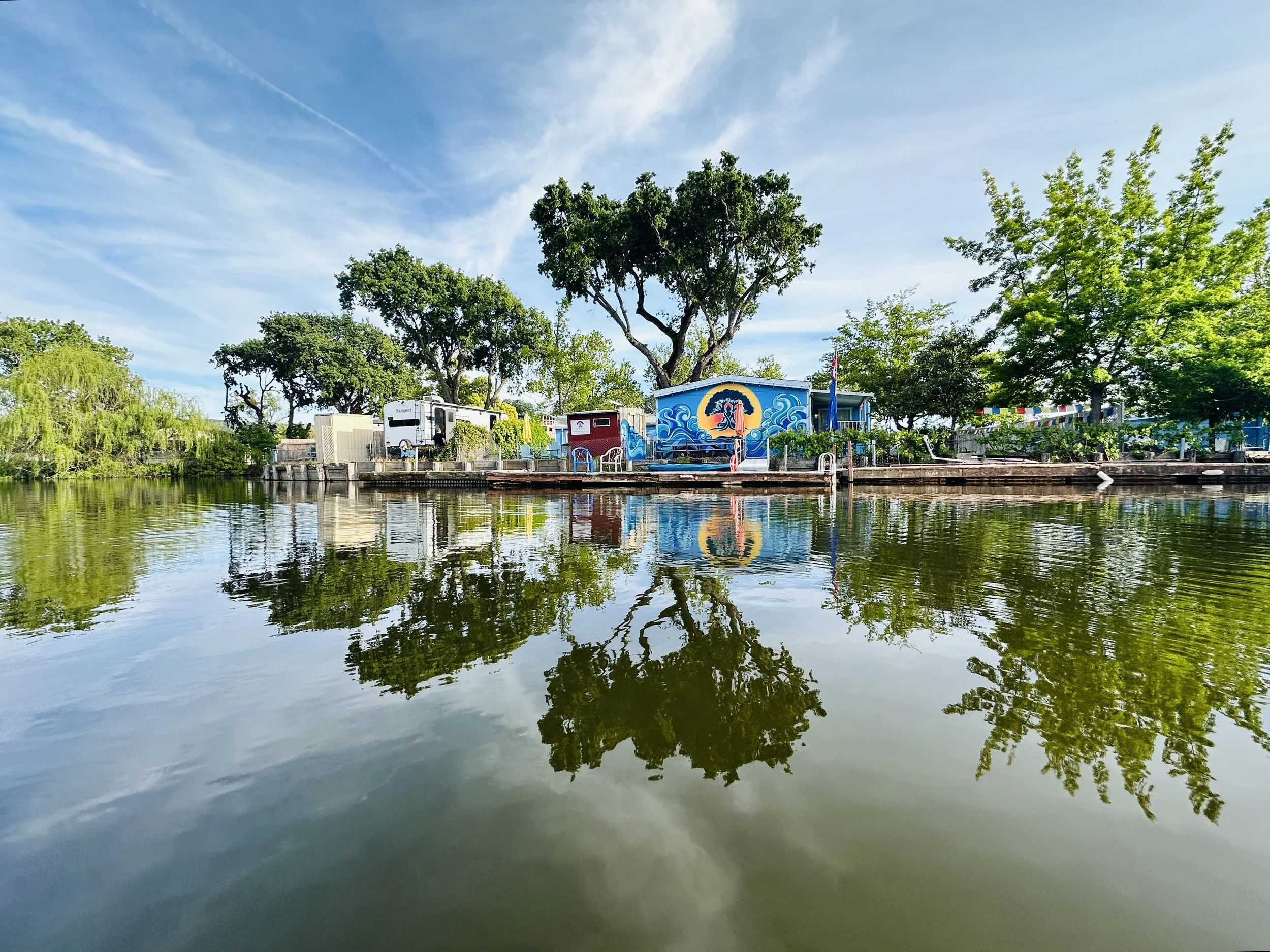 View of a small park or recreational area with a colorful mural on a blue building, surrounded by green trees, reflected on a calm body of water, under a partly cloudy sky.