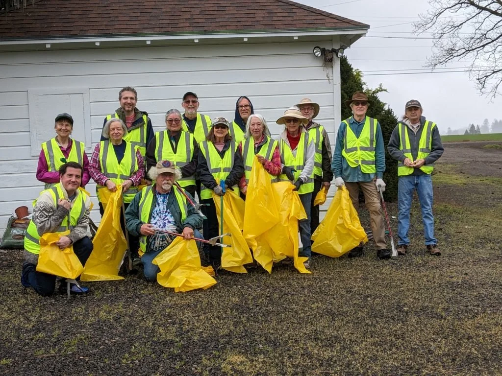 Group of volunteers wearing yellow safety vests and yellow trash bags, posing for a photo outdoors in front of a white building participating in a community cleanup effort.