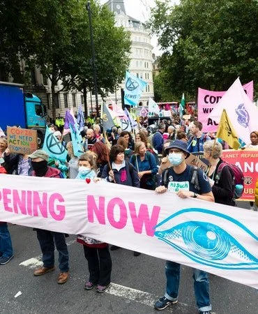 Group of people protesting on a city street, holding signs and banners about climate action, including a large banner with an eye symbol and the word "now."