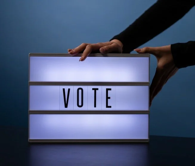 A person in a dark suit is adjusting a lit sign with the word "VOTE" on it