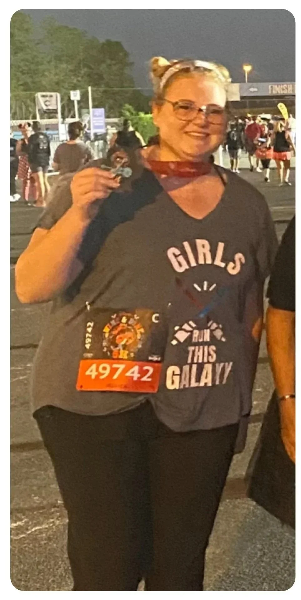 A woman smiling at a night marathon event, holding a medal with a race bib number 49742, wearing a black t-shirt with the words 'GIRLS RUN THIS GALAXY' and a badge with a lion emblem.