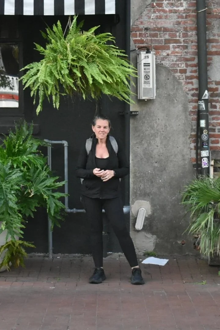 A woman standing on a brick sidewalk in front of a brick and concrete building, surrounded by green plants and ferns, with a hanging fern plant above her head.