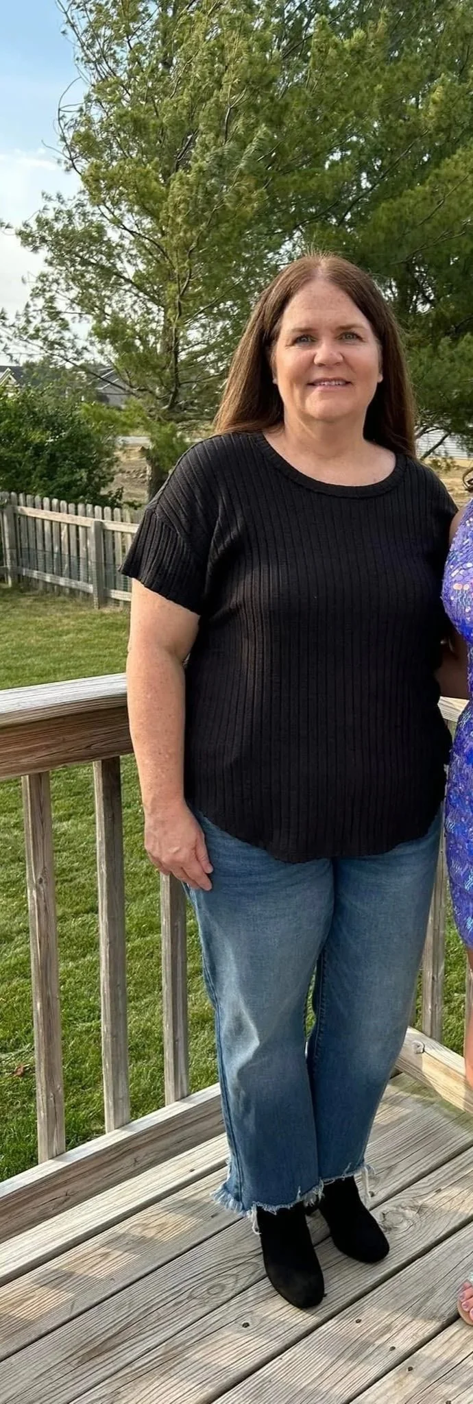 A woman standing on a wooden deck outdoors, wearing a black ribbed t-shirt, ripped jeans, and black boots. In the background, there are green trees, a wooden fence, and a partly cloudy sky.