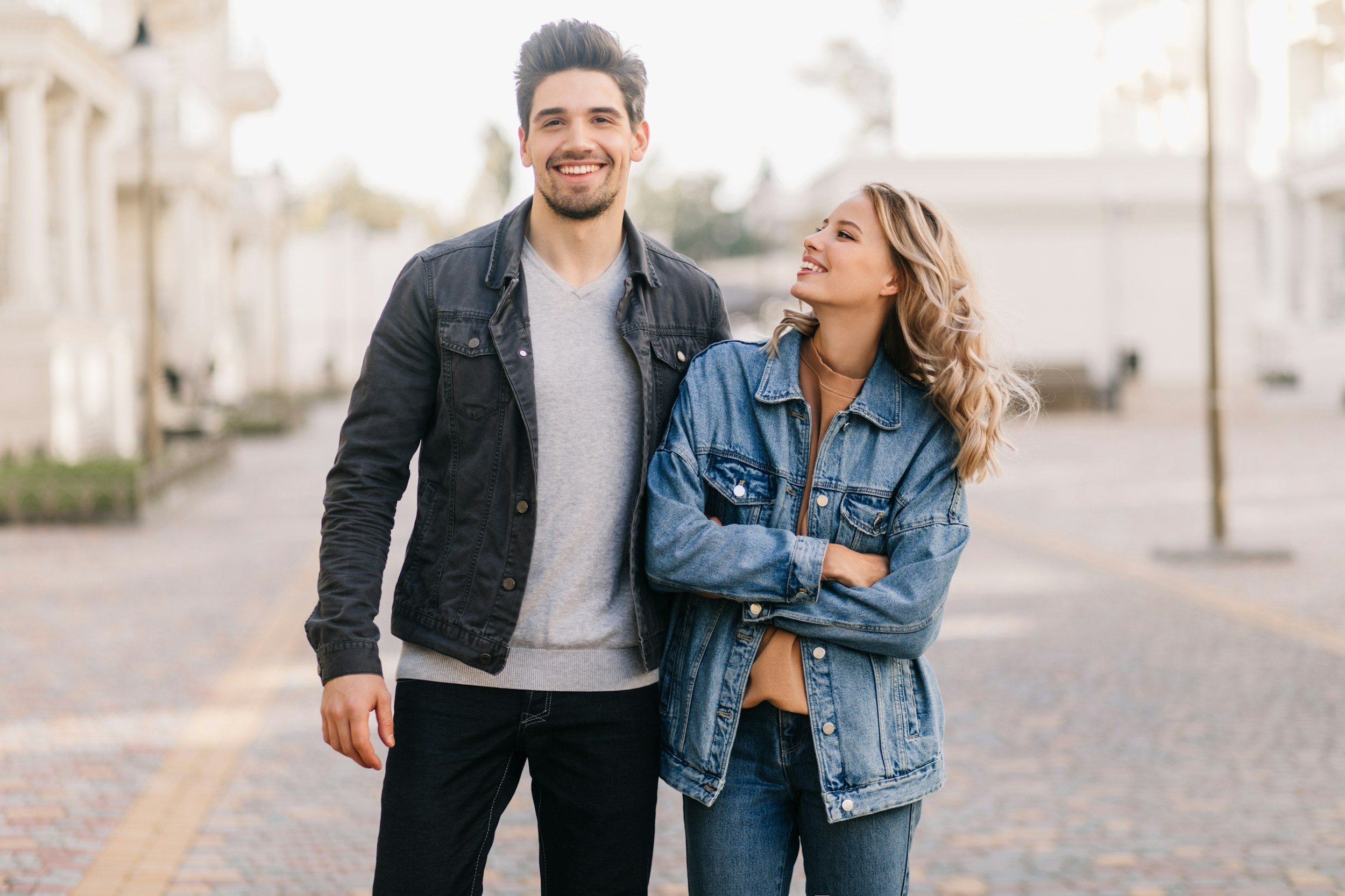 A young man and woman walking outdoors on a sunny day, smiling and looking happy. The man has dark hair and is wearing a denim jacket and gray t-shirt. The woman has long blonde hair and is wearing a denim jacket and tan-colored shirt. They are walking on a paved path with blurred buildings and trees in the background.