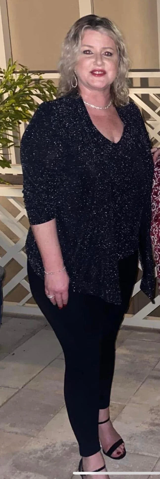 A woman with curly blonde hair wearing a sparkly black top, black pants, and black high heels, standing outdoors at night near a white lattice fence and a potted plant.