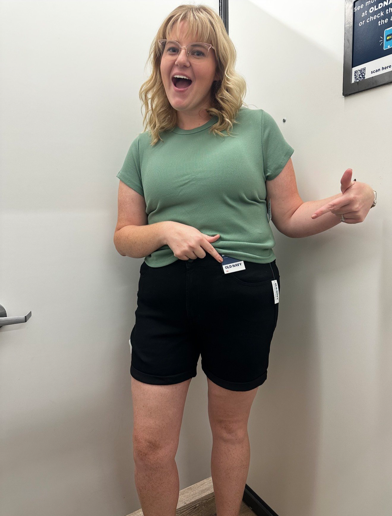 A woman with blonde curly hair, glasses, and a green t-shirt pointing to her waist with her right hand and smiling, standing in front of a white wall.
