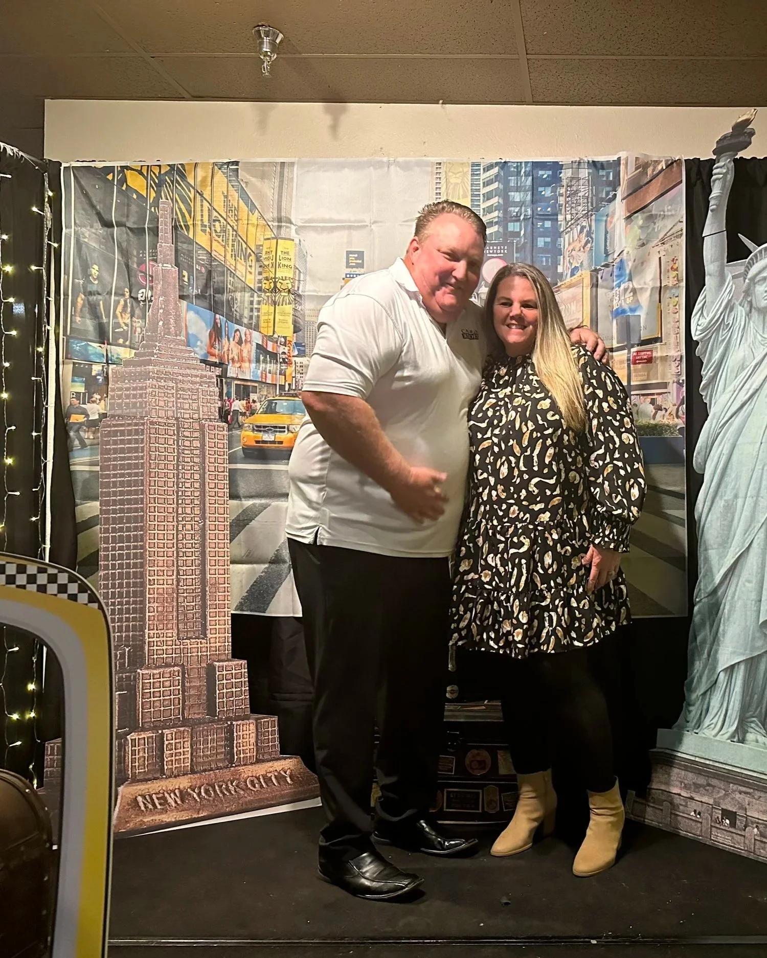 A man and a woman standing together smiling, with a backdrop of Times Square, New York City, showing buildings, taxis, and Billboards. The man wears a white T-shirt and black pants, and the woman wears a black dress with a patterned design and beige boots.