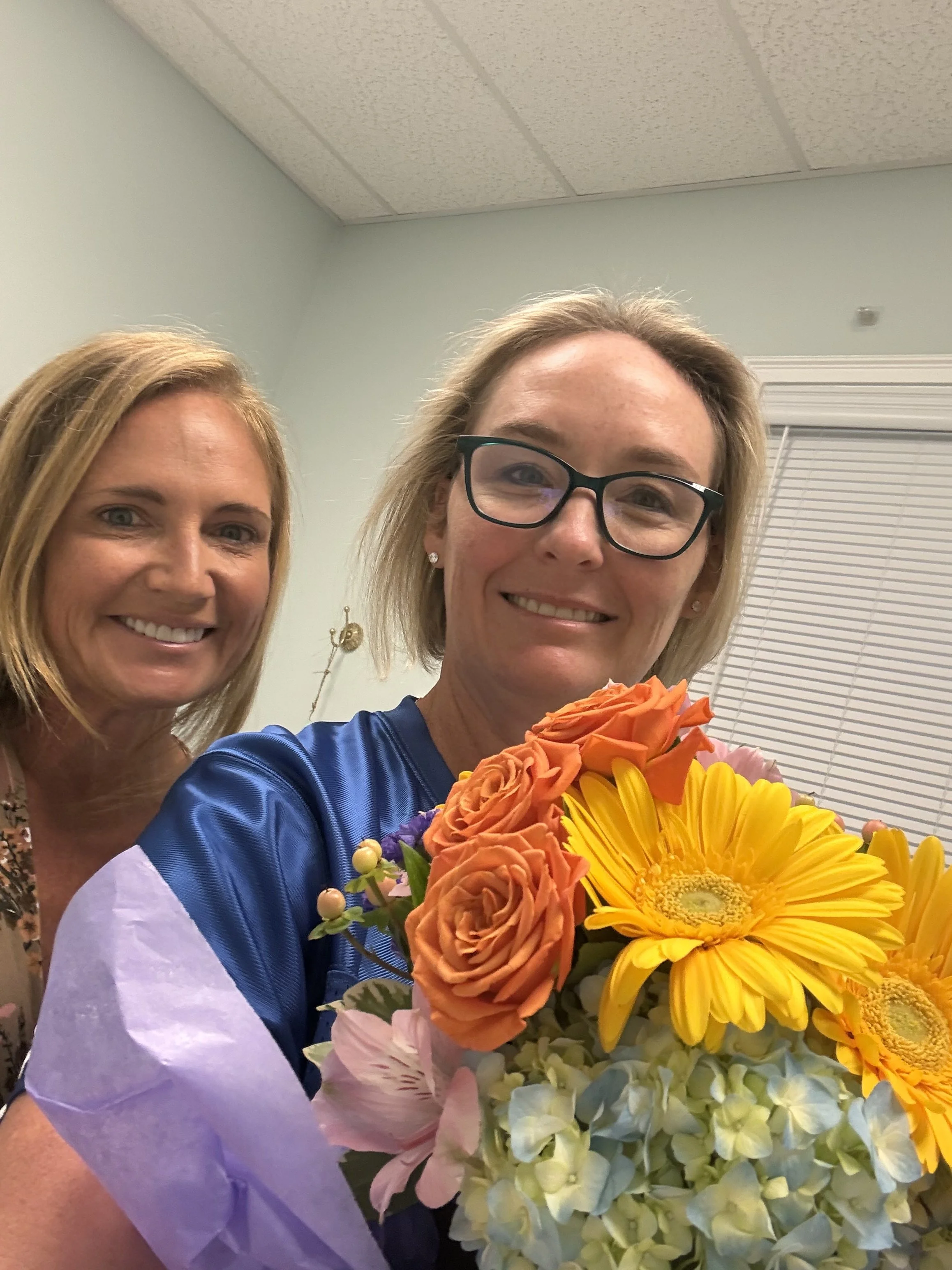 client and Nurse practitioner Lisa Sikorski -Two women, one with blonde hair and glasses holding a colorful bouquet of flowers, smiling in an indoor setting with closed blinds.