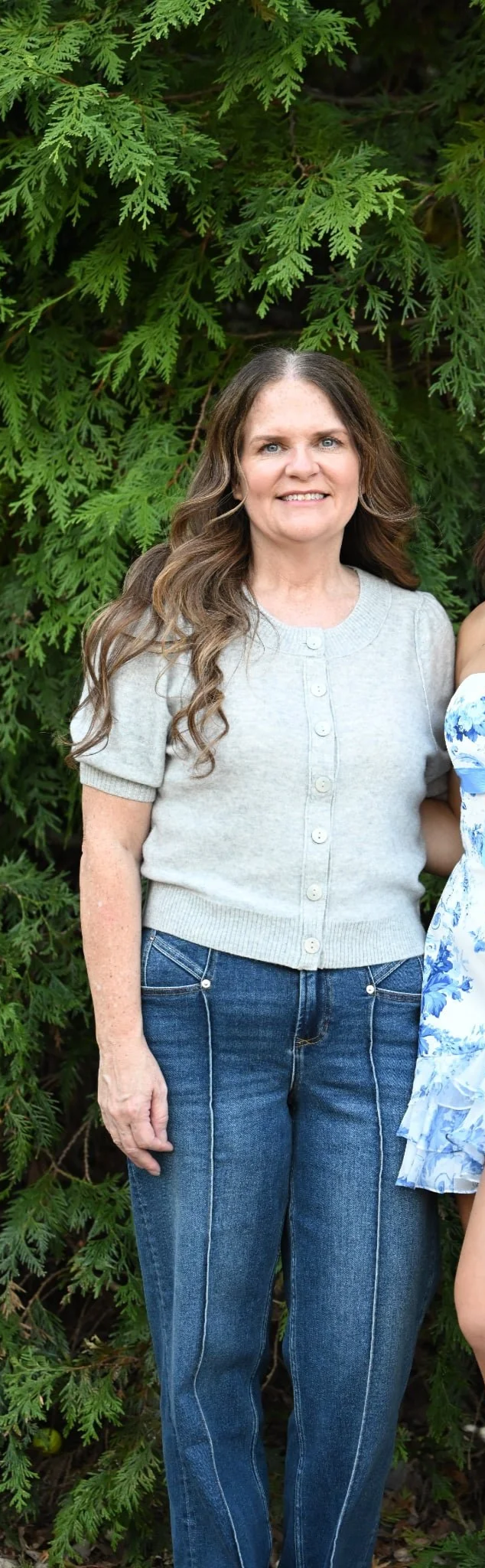 A woman with long wavy brown hair and blue eyes smiling, wearing a light gray short-sleeved sweater and blue jeans, standing outdoors in front of lush green foliage.