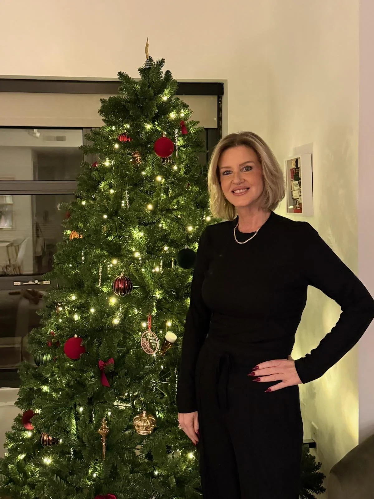 A woman with blonde hair dressed in black standing next to a decorated Christmas tree with ornaments and lights.