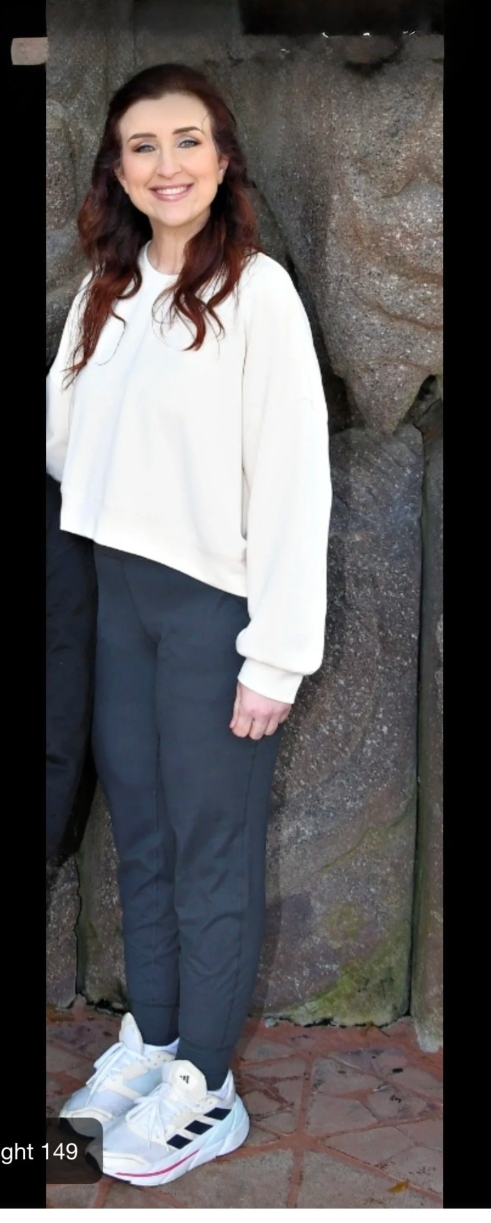 A woman with long red hair smiling, dressed in a white sweater, black pants, and white sneakers with black stripes, standing against a stone wall with brick ground.