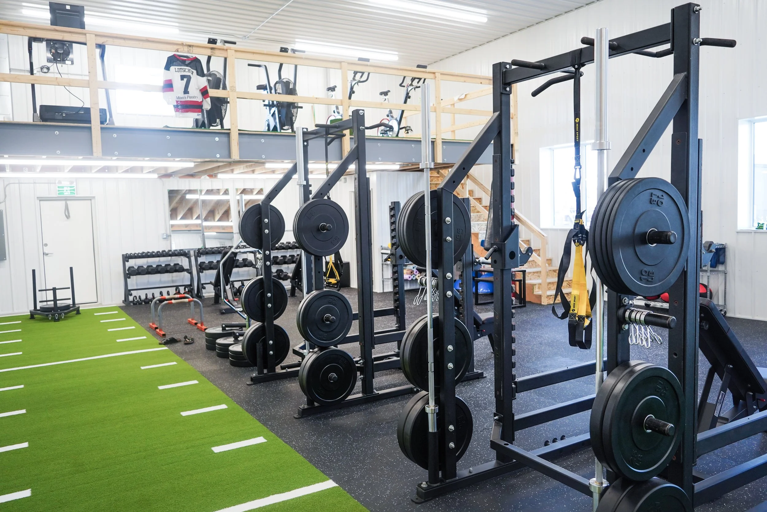 Indoor gym with weightlifting equipment, including barbells, weight plates, and machines. There is a green artificial turf zone on the floor, a wooden loft with bikes and hockey jersey and a staircase leading to the upper level. Mitch Dyck Perfomance