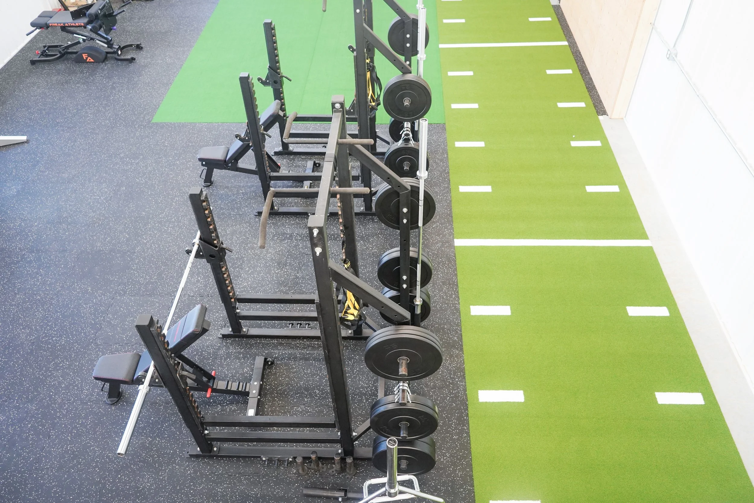 Weightlifting gloves, barbells, weight plates, and athletic equipment in a gym with green flooring and black rubber mats. PV Elite Hockey, Mitch Dyck Performance Centre
