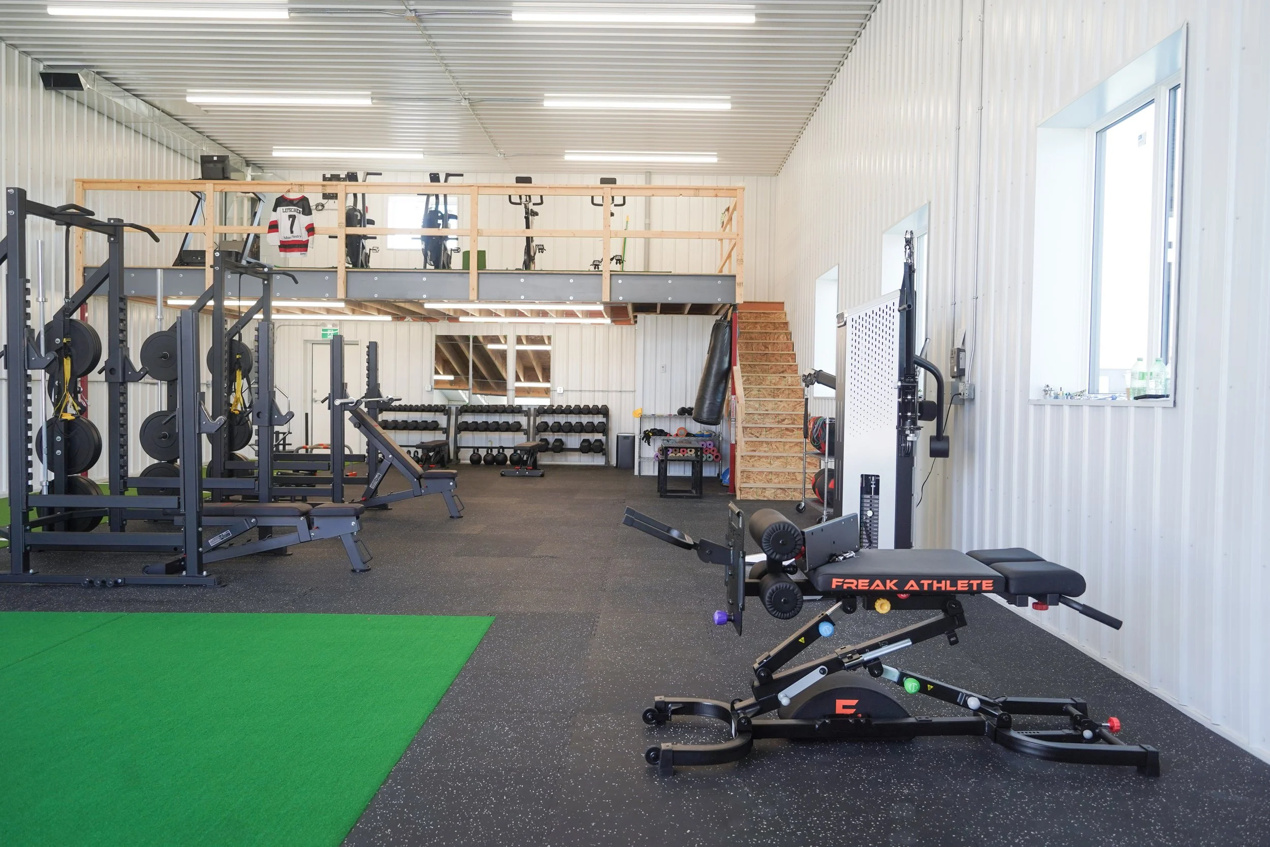 Interior of a gym with workout equipment including squat rack, weights, resistance bands, punching bag, and exercise bike, with a small loft area above. Off-season hockey program hockey training facility hockey training