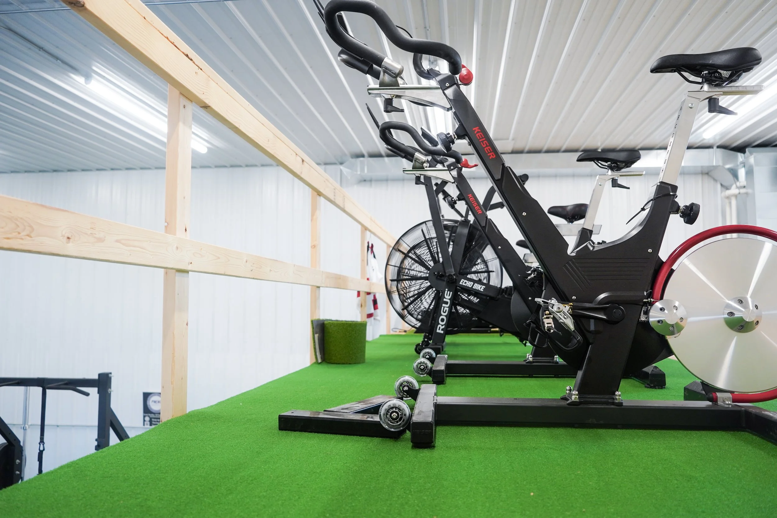 Indoor gym featuring a black Keiser exercise bike on green turf flooring, with dim lighting and a wooden railing alongside the workout area. Off-season hockey training, PV Elite Hockey, Mitch Dyck Performance Centre