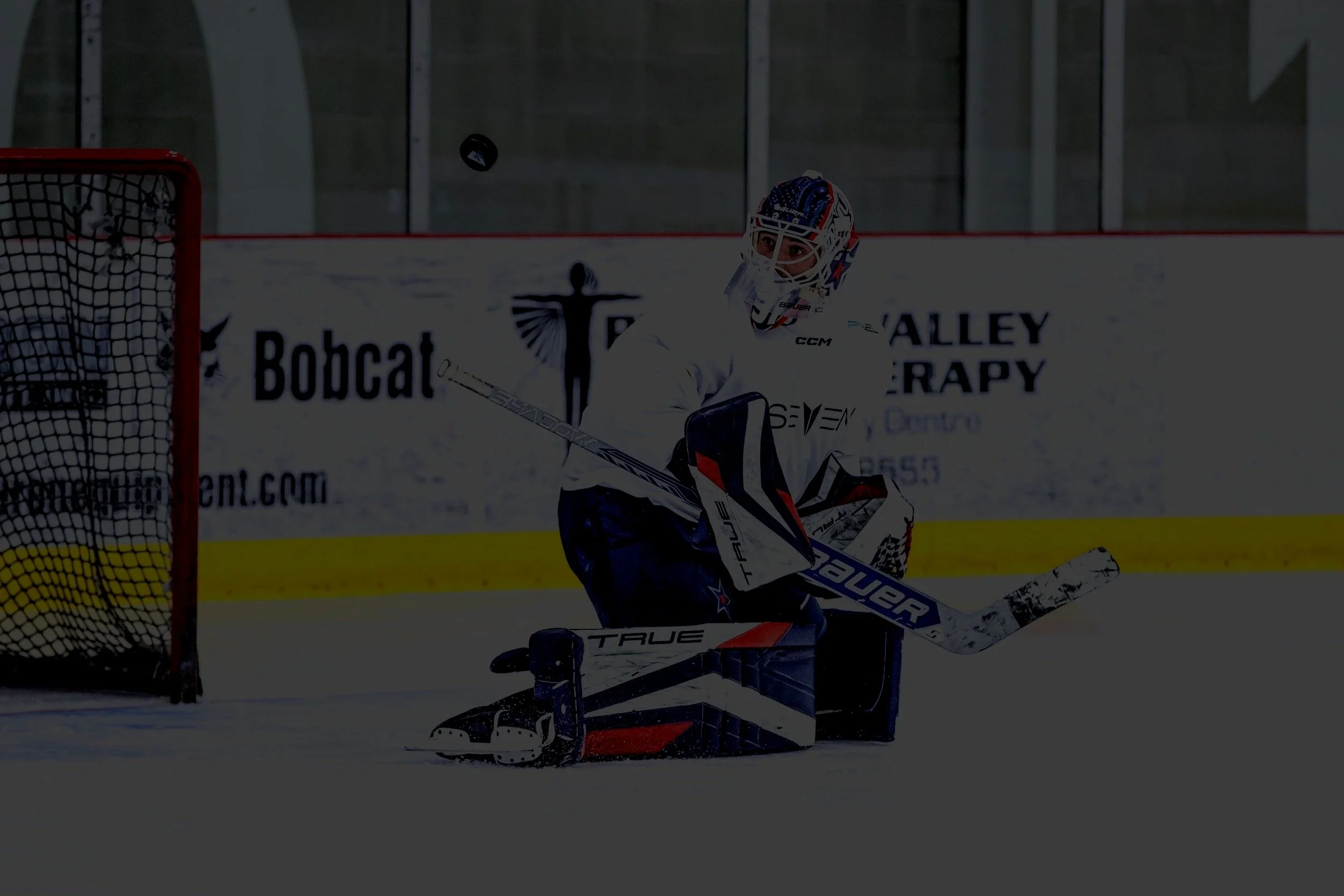 A hockey goalie in white and blue gear is on the ice, ready to make a save, with a black puck in midair near the net. Goaltending Development PV Elite Hockey.