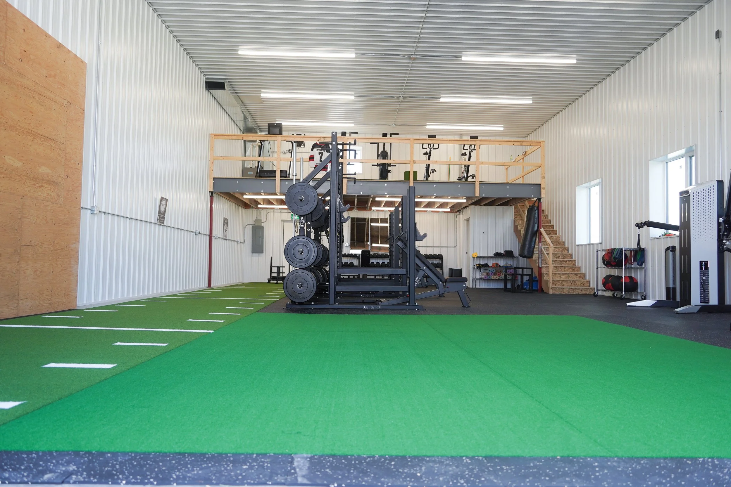 Empty gym with green turf, workout equipment, and a lofted area with more exercise machines and a staircase. PV Elite Hockey, Mitch Dyck Performance Centre