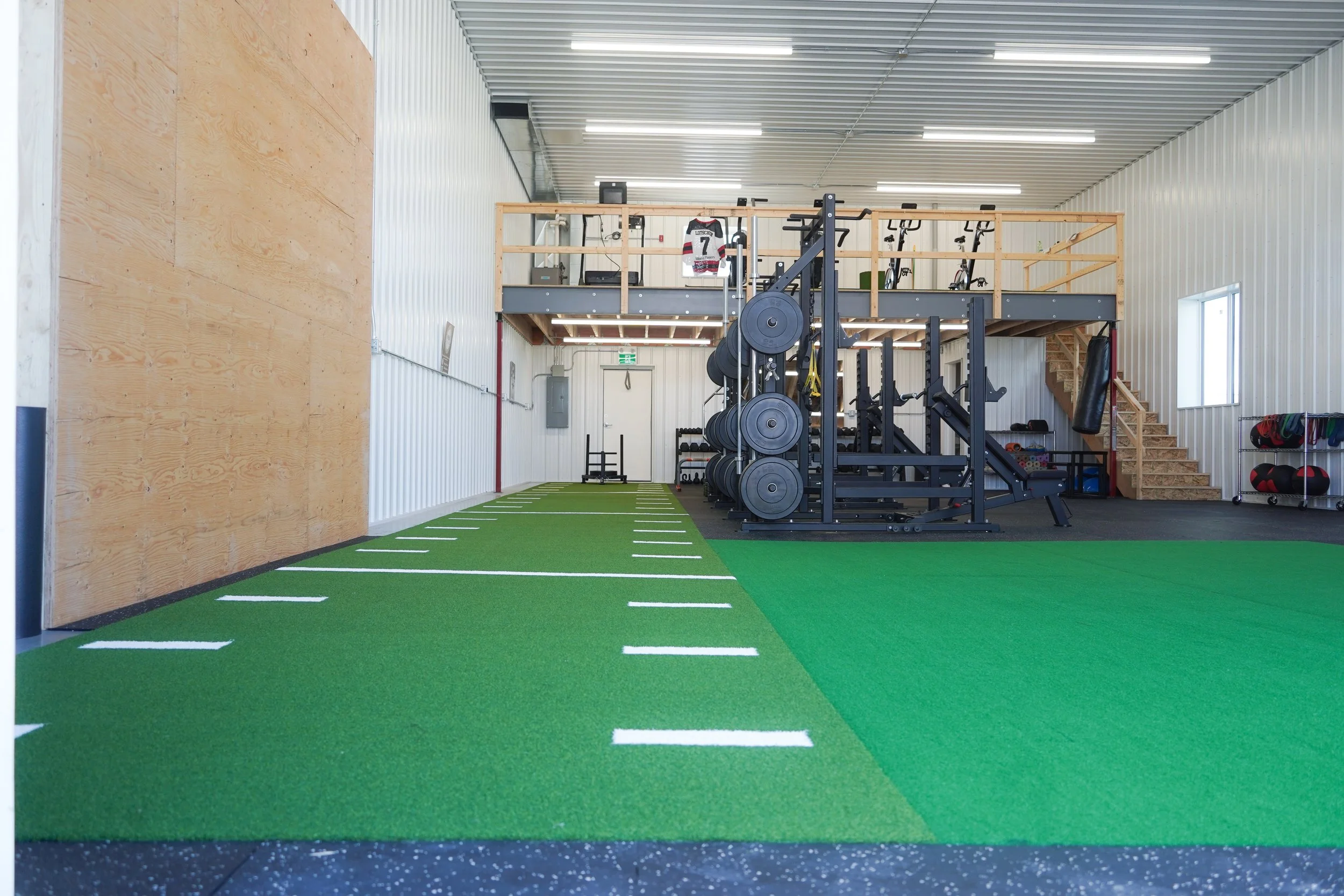 Empty workout gym with artificial turf parking lines on the floor, weightlifting equipment including barbells and weights, resistance machines, and a mezzanine area with jerseys and bikes, all inside a spacious, well-lit steel-paneled building Hockey