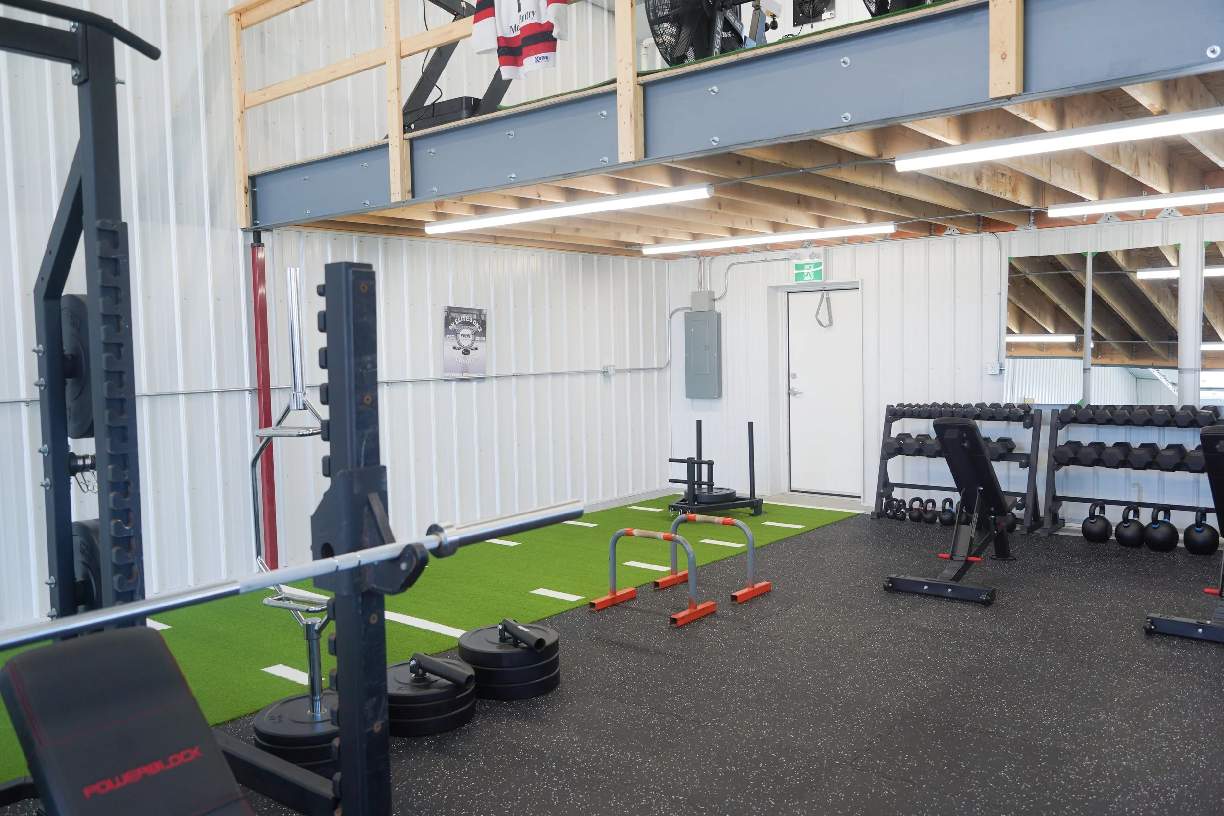 Interior of a gym with various workout equipment including weights, kettlebells, and exercise machines, with a mezzanine level above. PV Elite Hockey, Mitch Dyck Performance Centre. Hockey Specific Training Facility