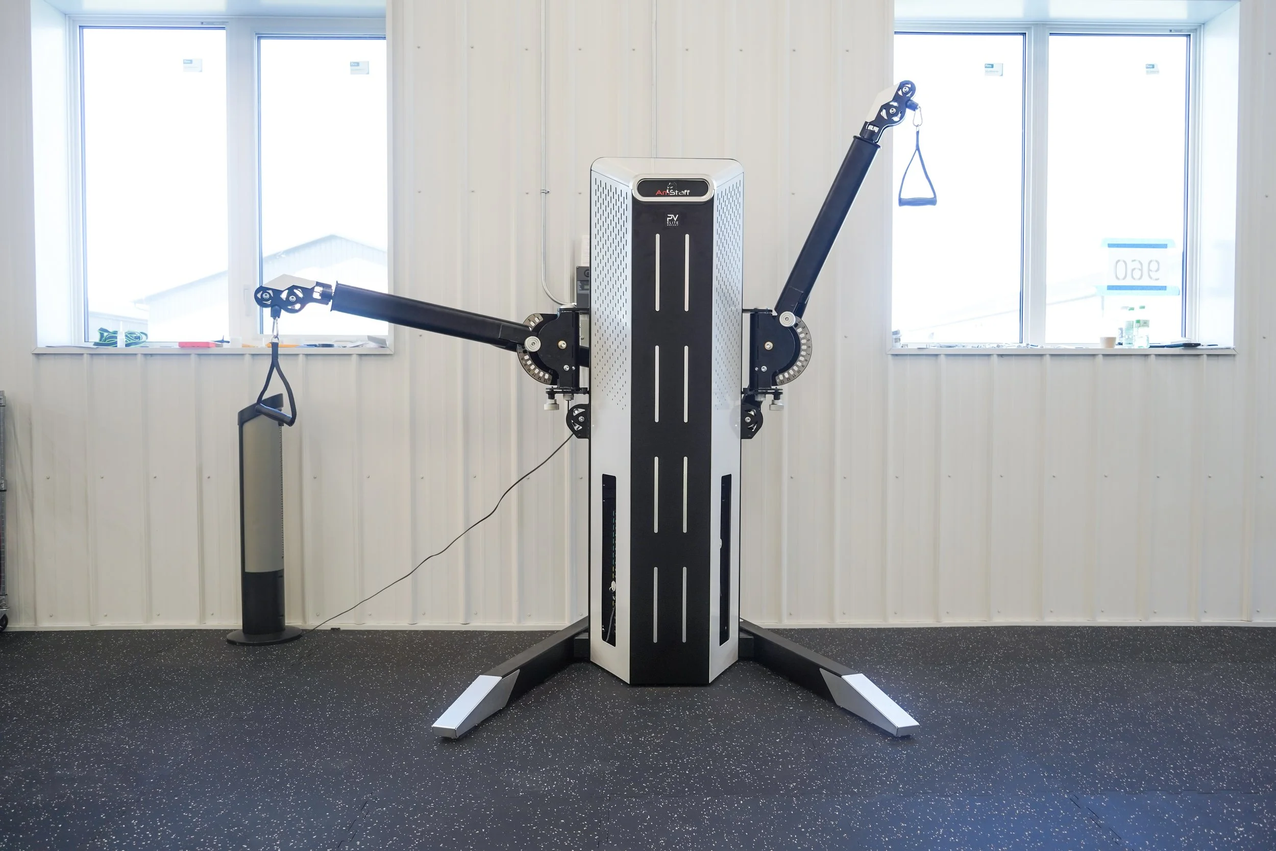 Robotic exercise equipment with two adjustable arms extending outward, each ending with handles, situated in a room with windows and a speckled black flooring. PV Elite Hockey, Mitch Dyck Performance Centre