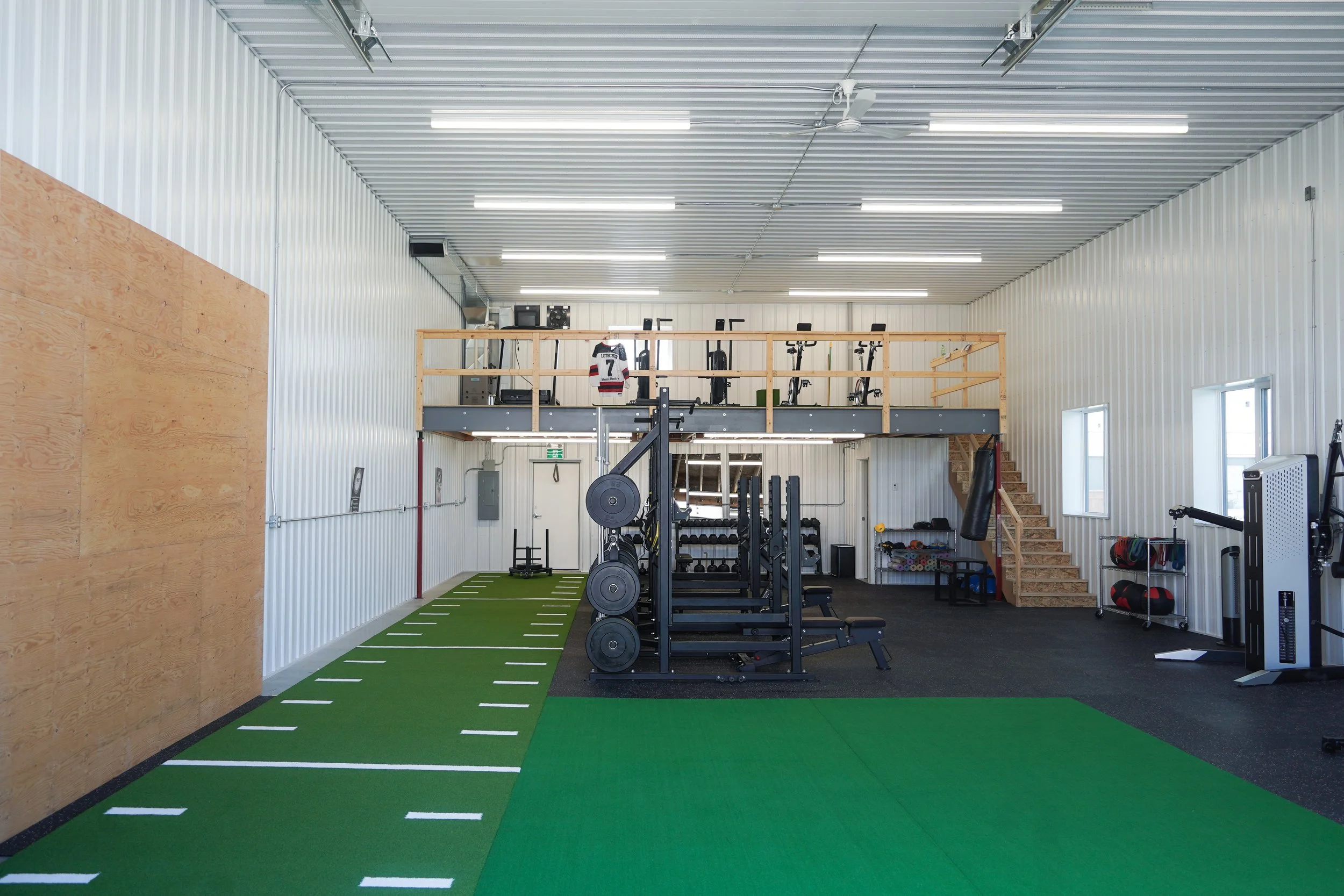 Inside a gym with weightlifting equipment, a green turf area, a mezzanine with exercise bikes, and wooden stairs leading up to the upper level. PV Elite Hockey, Mitch Dyck Performance Centre. Hockey Specific Training, Hockey Training