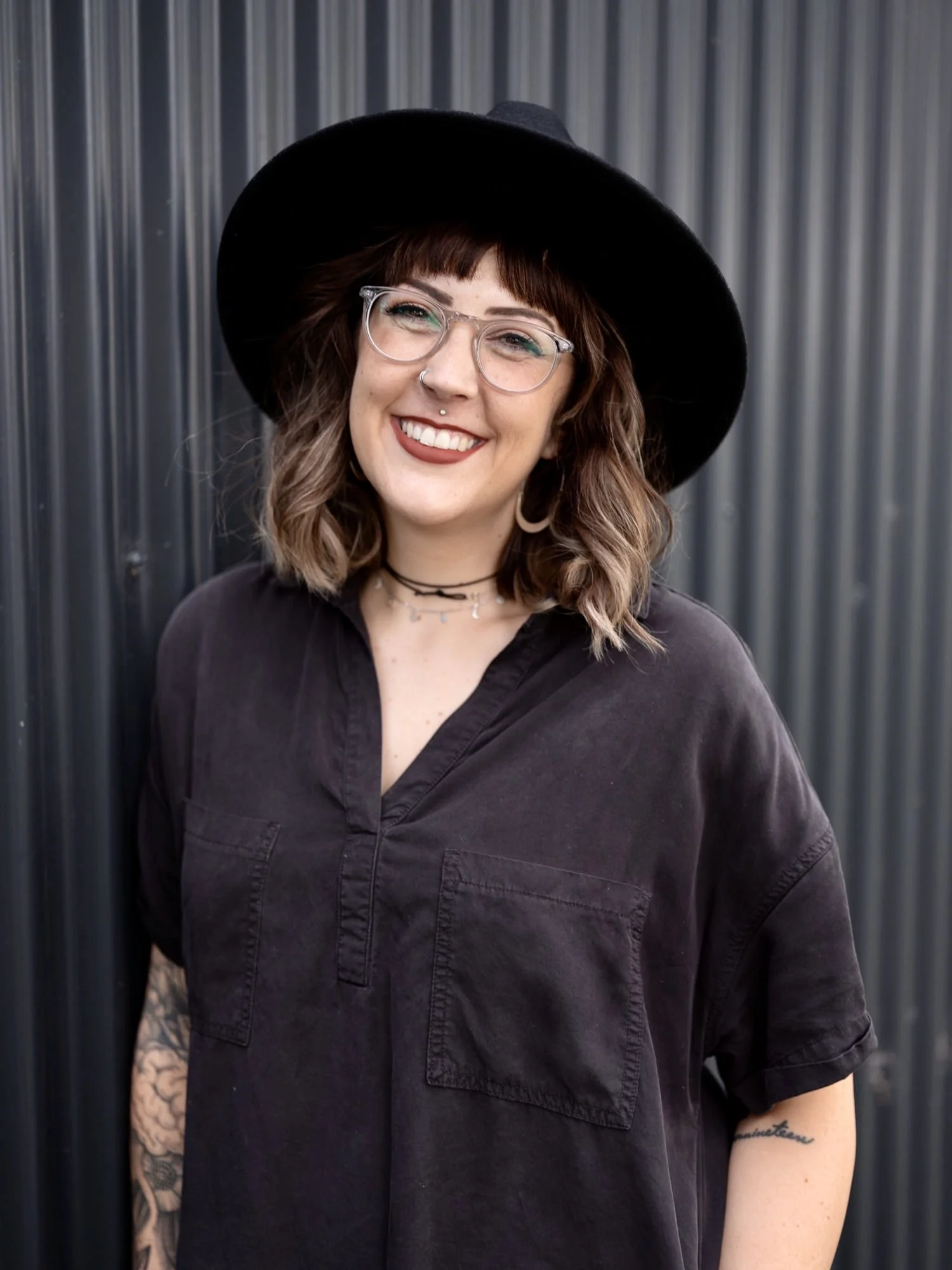 A smiling woman with glasses, tattoos on her arm, wearing a black hat and dark shirt, standing in front of a metal wall.