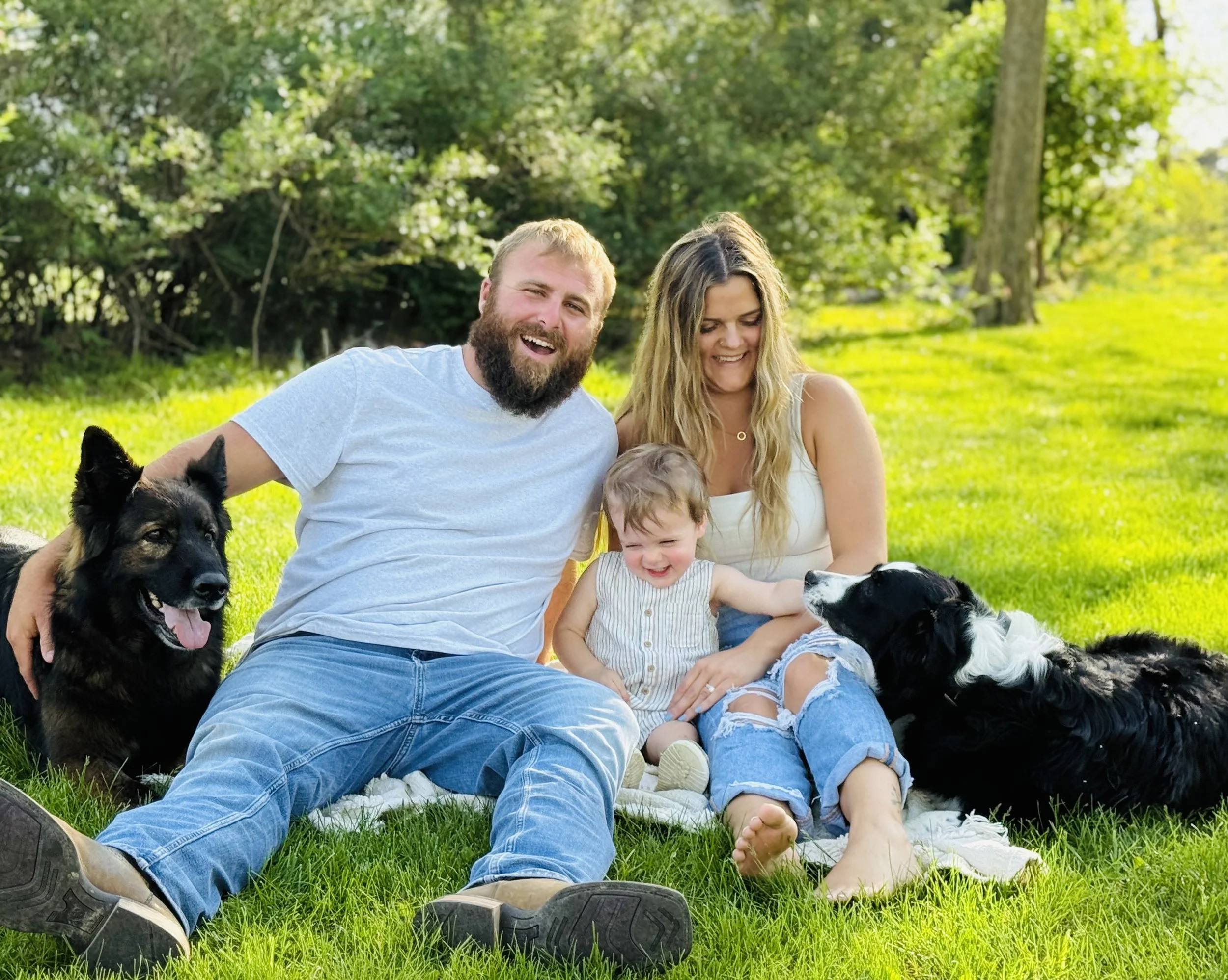 A happy family of four with two dogs sitting on grass in a park on a sunny day.