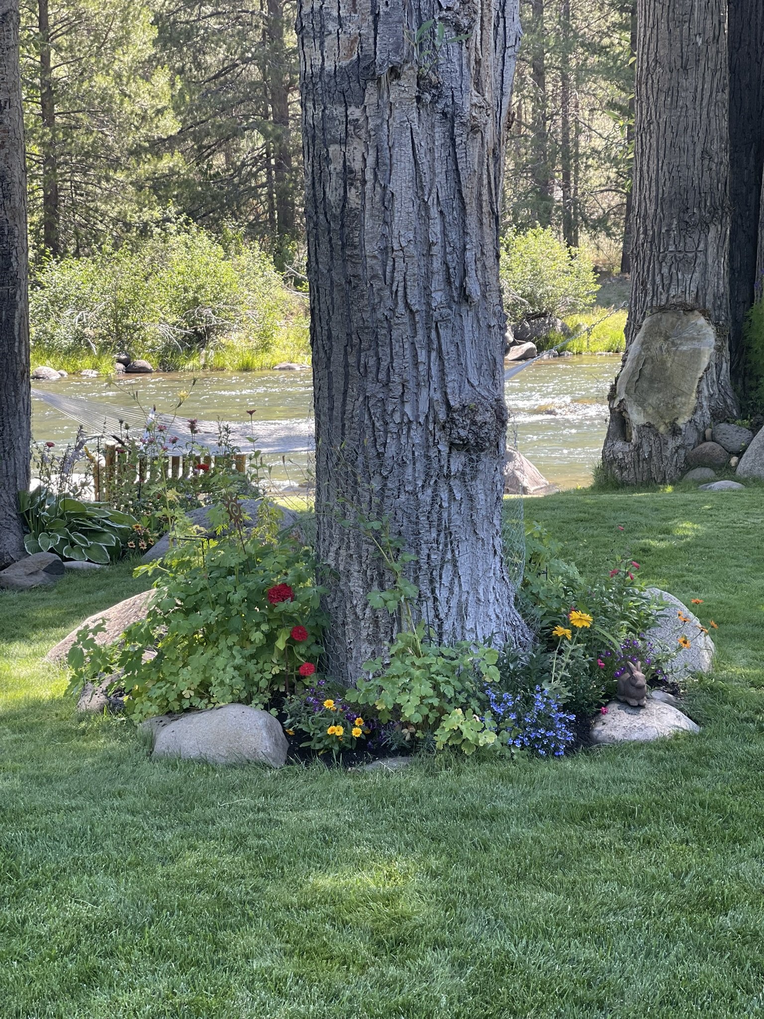 A garden scene with two large pine trees, a small river or stream in the background, and a colorful flower bed with rocks and a small garden ornament in the foreground. The flowers are yellow, red, purple, and blue.