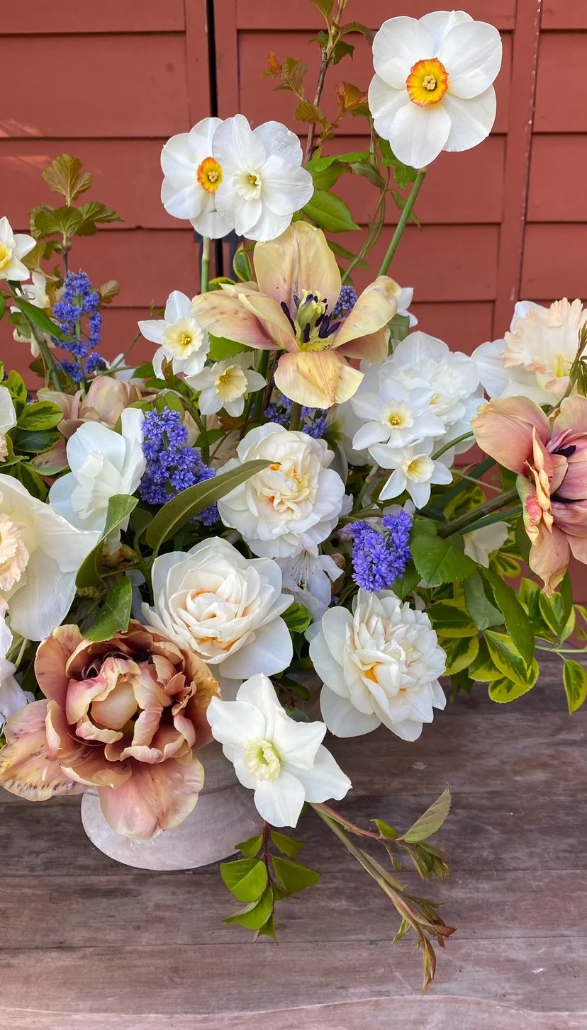 A colorful bouquet of various flowers, including white pansies, light pink tulips, and purple flowers, arranged in a white vase on a wooden surface with a reddish-brown wooden fence in the background.