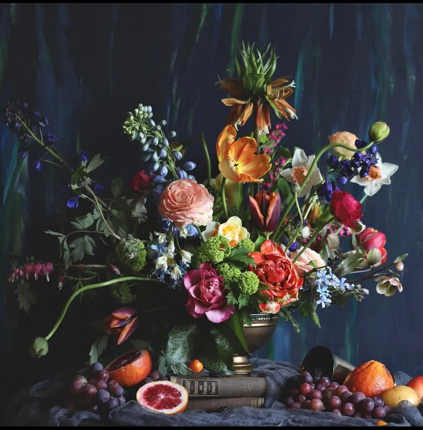 A colorful floral arrangement with various flowers, fruits, and greenery in a decorative vase, set against a dark backdrop.