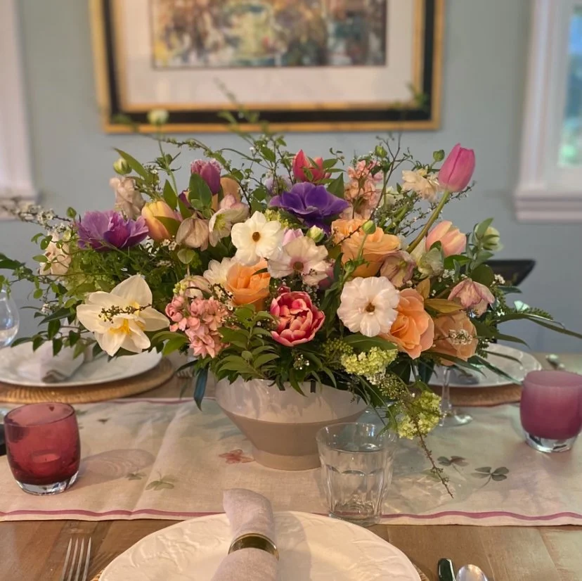 A floral centerpiece with pink, purple, white, and peach flowers on a dining table, surrounded by pink and clear glasses, and place settings with napkins and silverware.