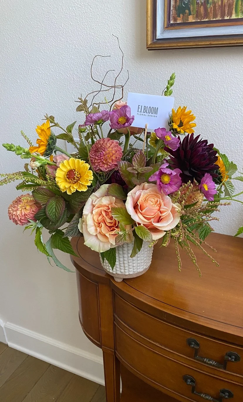 Colorful flower arrangement in a white textured vase on a wooden table, with a business card reading 'F.J. Bloom Floral Design' among the flowers.