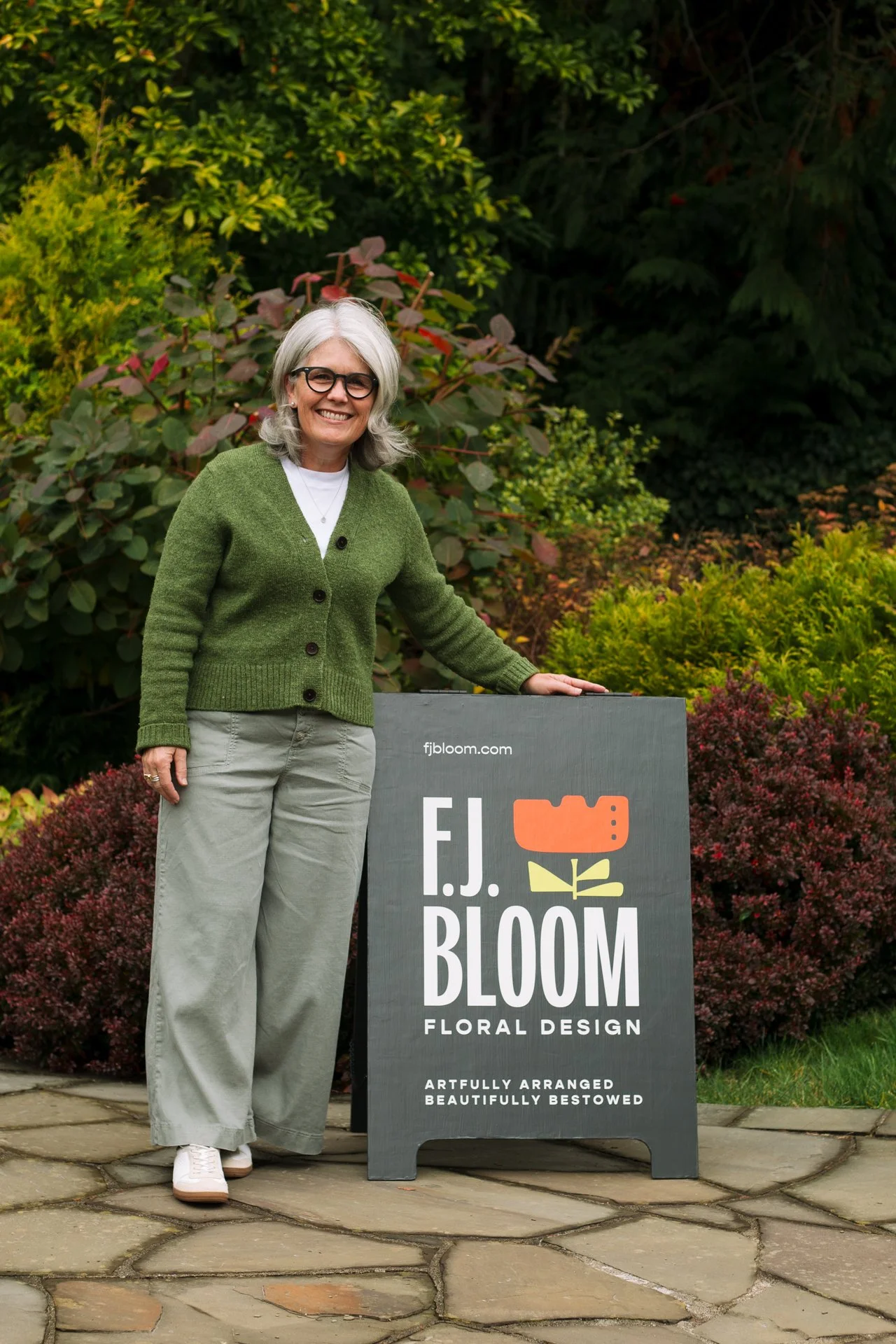 A woman with gray hair, glasses, wearing a green cardigan, standing outdoors next to a sign for F.J. Bloom Floral Design, surrounded by greenery and bushes.