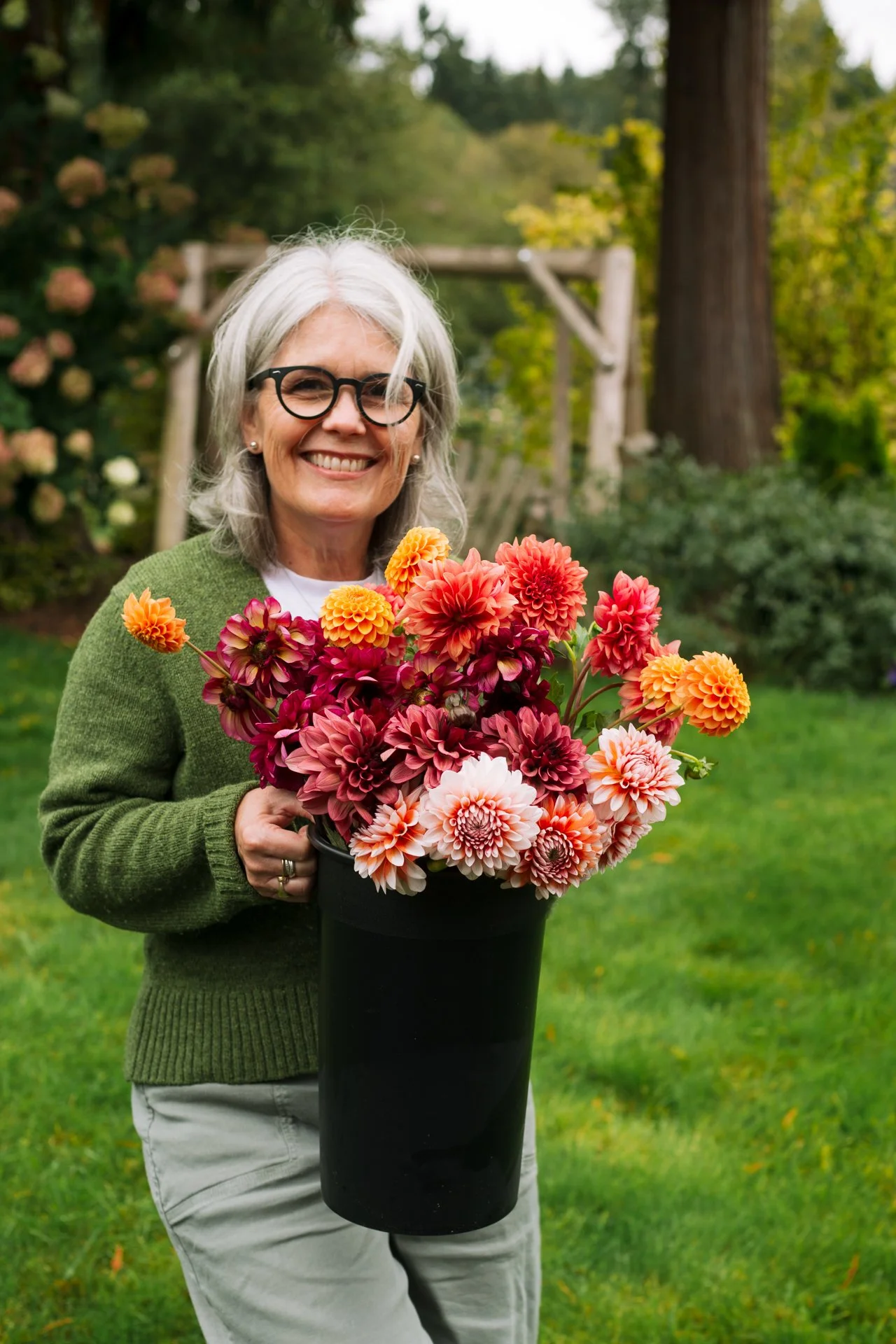 A smiling woman with gray hair, glasses, and pearl earrings holding a large bouquet of colorful dahlias in a black vase outdoors in a garden.