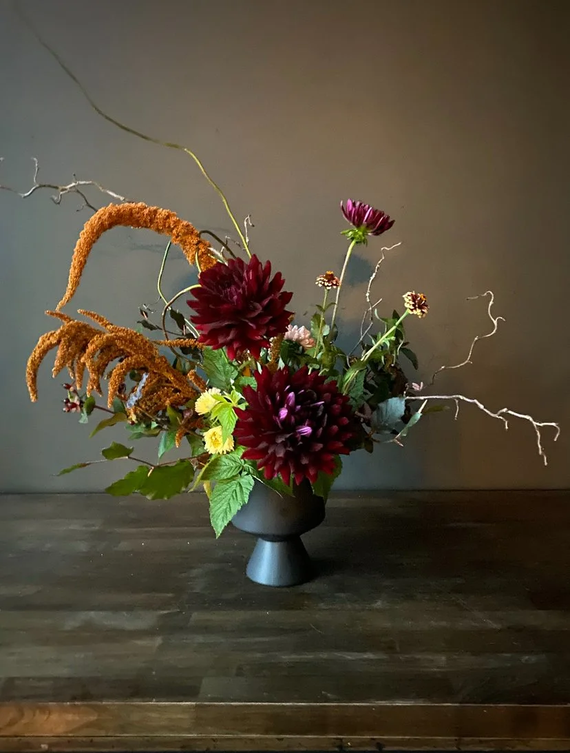 A floral arrangement with dark red flowers, green leaves, and dried branches in a black vase on a wooden table against a gray wall.