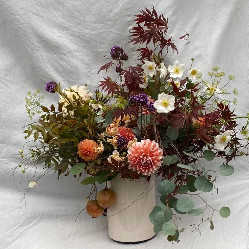 A floral arrangement in a white vase with blush pink dahlias, white flowers, purple and yellow accents, reddish leaves, and green foliage against a crinkled light gray background.