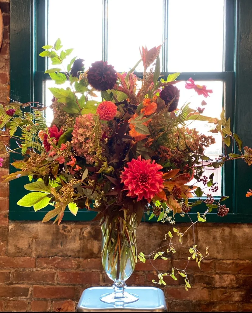 A large bouquet of flowers in a clear glass vase on a small table in front of a window with green frames, against a brick wall.