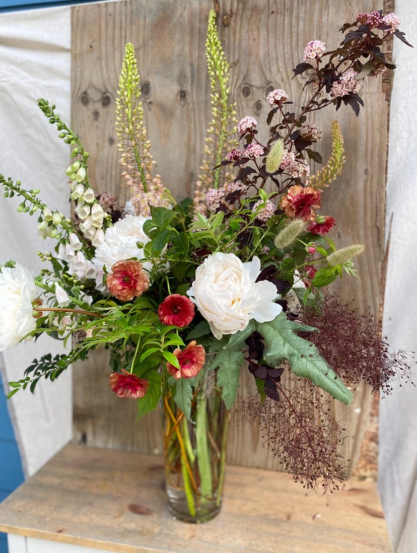 A glass vase filled with a mixed flower bouquet including white roses, red and peach-colored flowers, green foliage, and tall purple and yellow flowering stems, placed on a wooden surface against a wooden panel background.