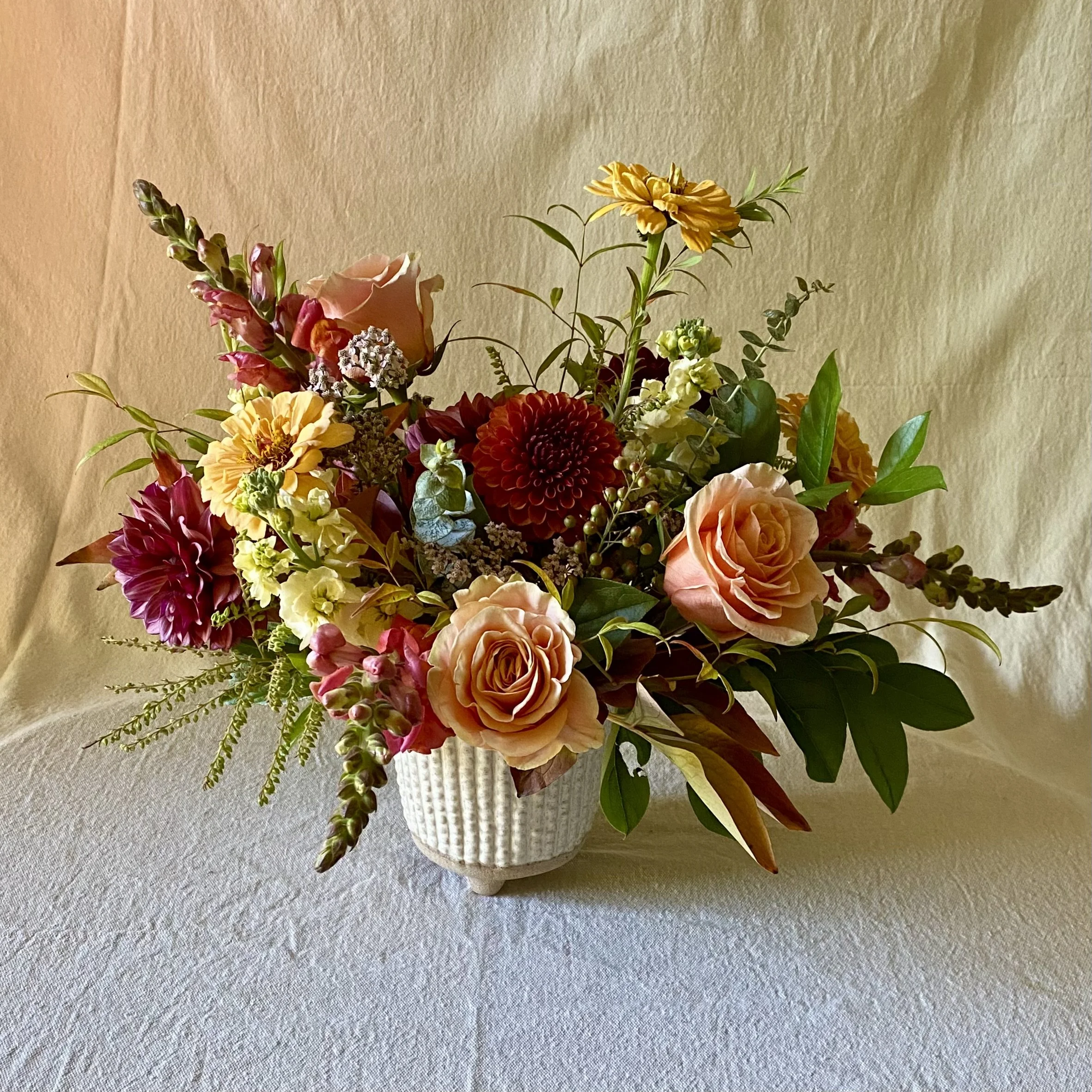 A flower arrangement in a white textured vase featuring pink roses, red dahlias, yellow and pink snapdragons, and various green foliage, set against a beige background.
