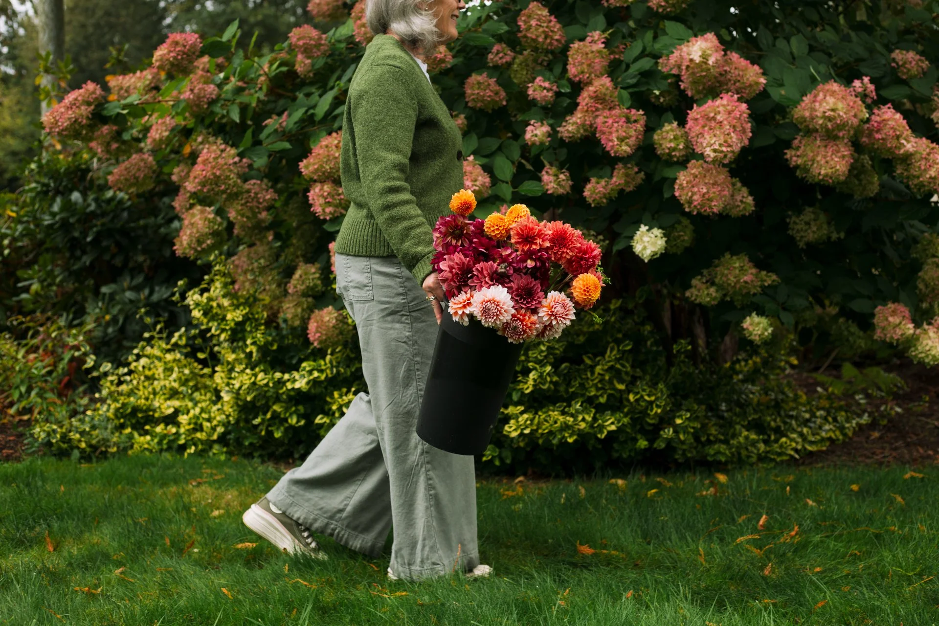 A woman with gray hair, wearing a green sweater and gray pants, standing outdoors on grass, holding a tall black vase filled with pink and orange flowers, with a bush of pink and green foliage in the background.
