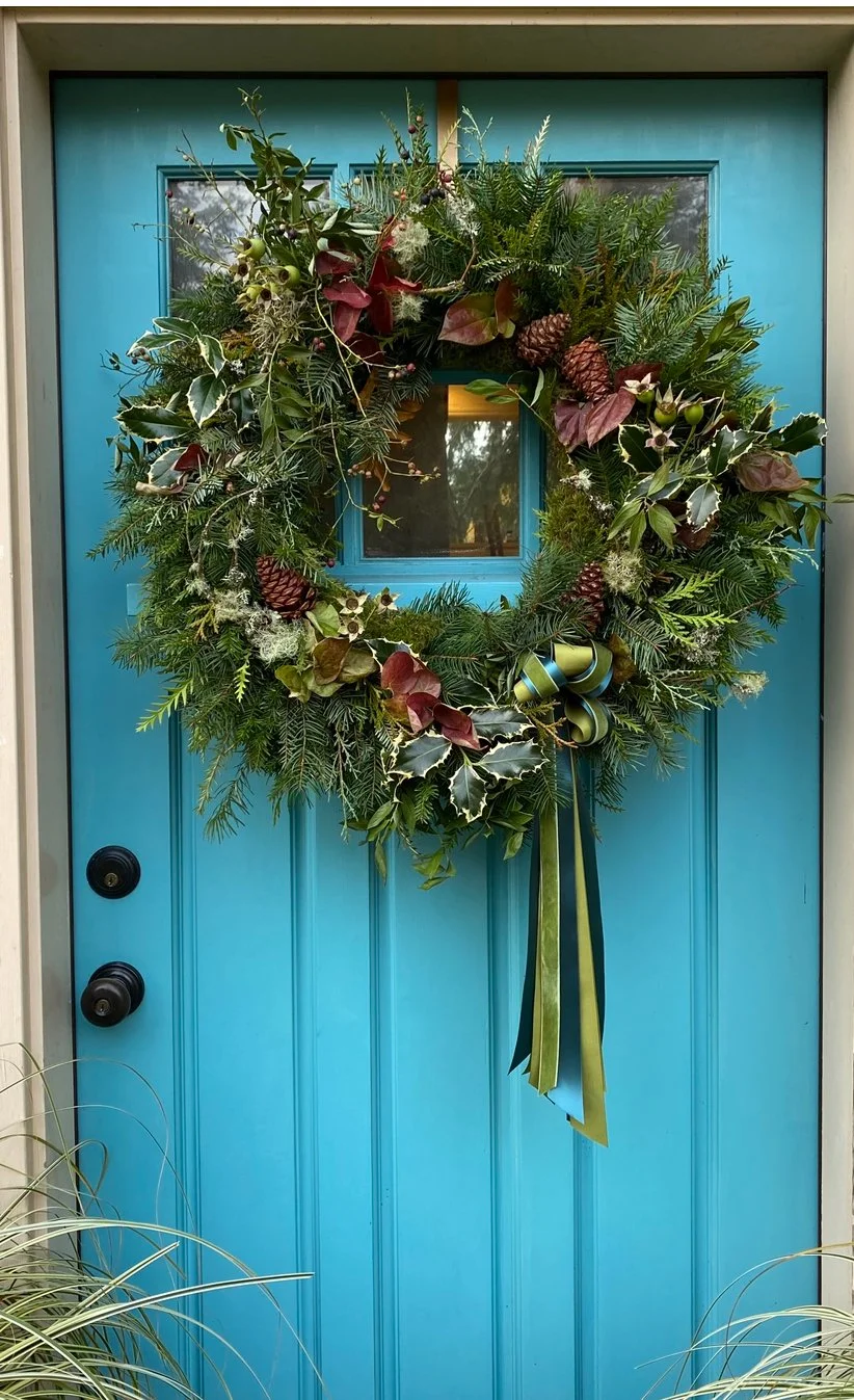A Christmas or holiday wreath hanging on a bright blue door. The wreath is made of assorted evergreen branches, holly leaves, pinecones, and red and green foliage, decorated with a green and gold ribbon.