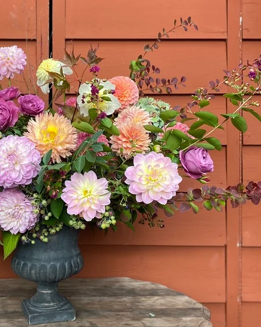 A floral arrangement in a dark gray vase featuring pink, purple, cream, and peach-colored flowers, with green foliage, set on a wooden surface against a red wooden fence background.