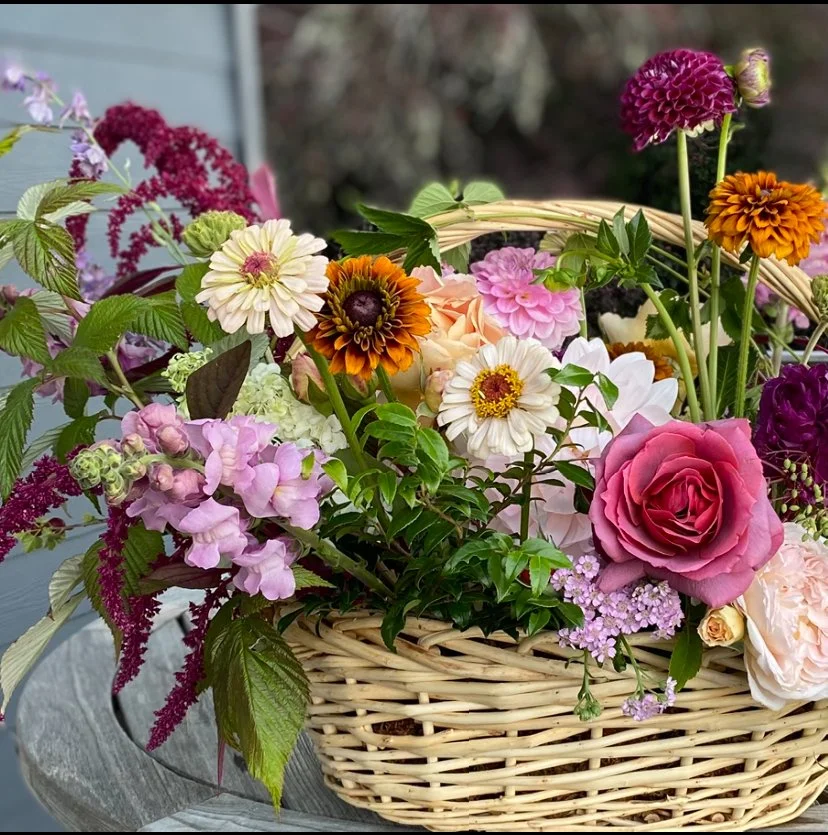 A wicker basket filled with a variety of colorful flowers including roses, dahlias, and other blossoms in pink, purple, orange, white, and yellow, placed on a gray surface outdoors.