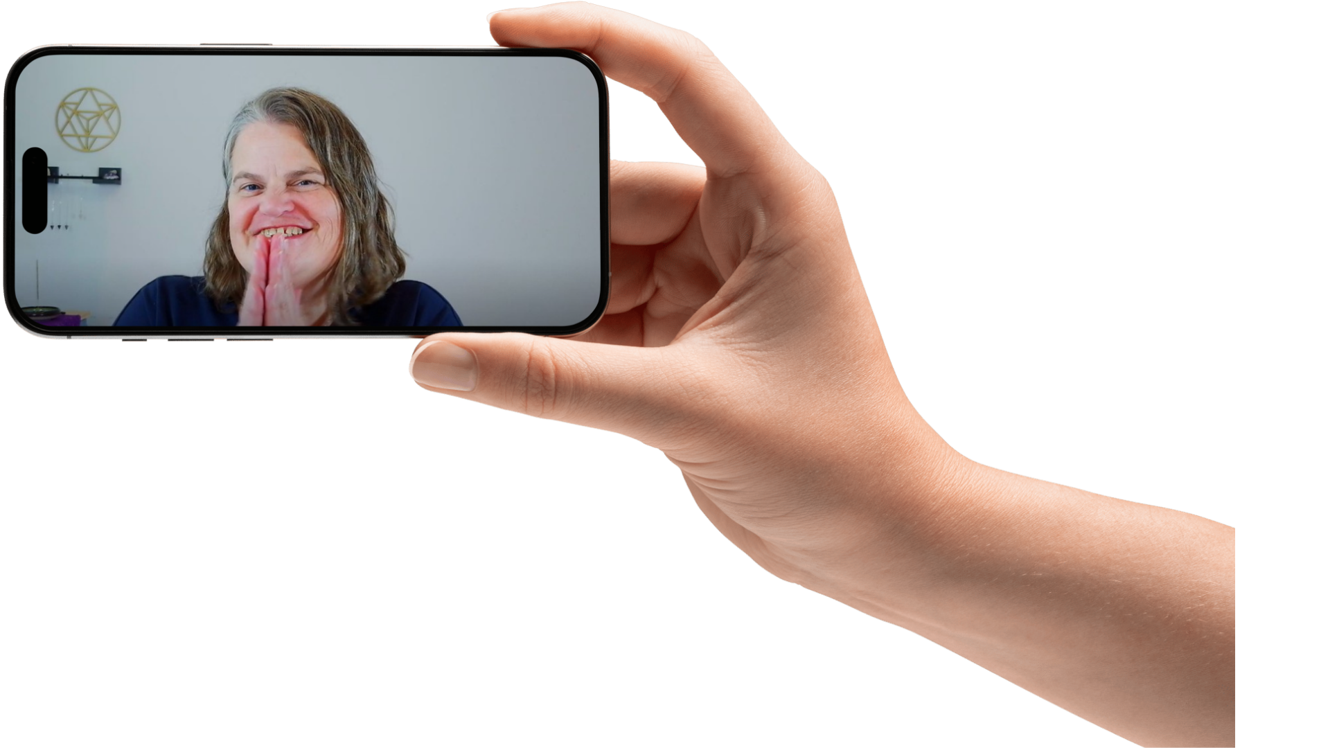 A person holding a smartphone taking a selfie of a smiling woman with short gray hair, clasping her hands in a prayer-like gesture, in a room with a white wall and decorative symbols.