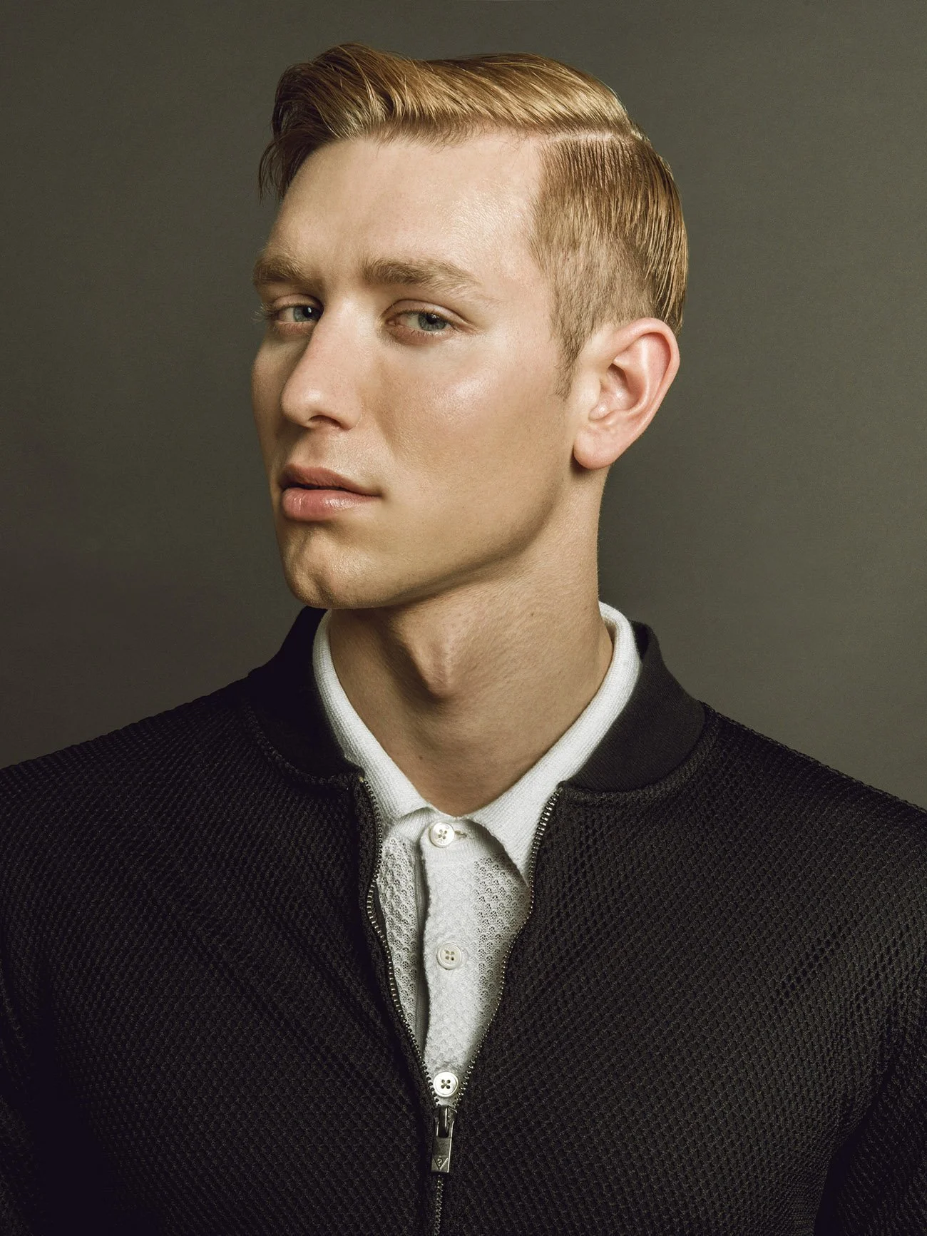 Portrait of a young man with blonde hair, wearing a black zip-up jacket over a white collared shirt, against a plain background.