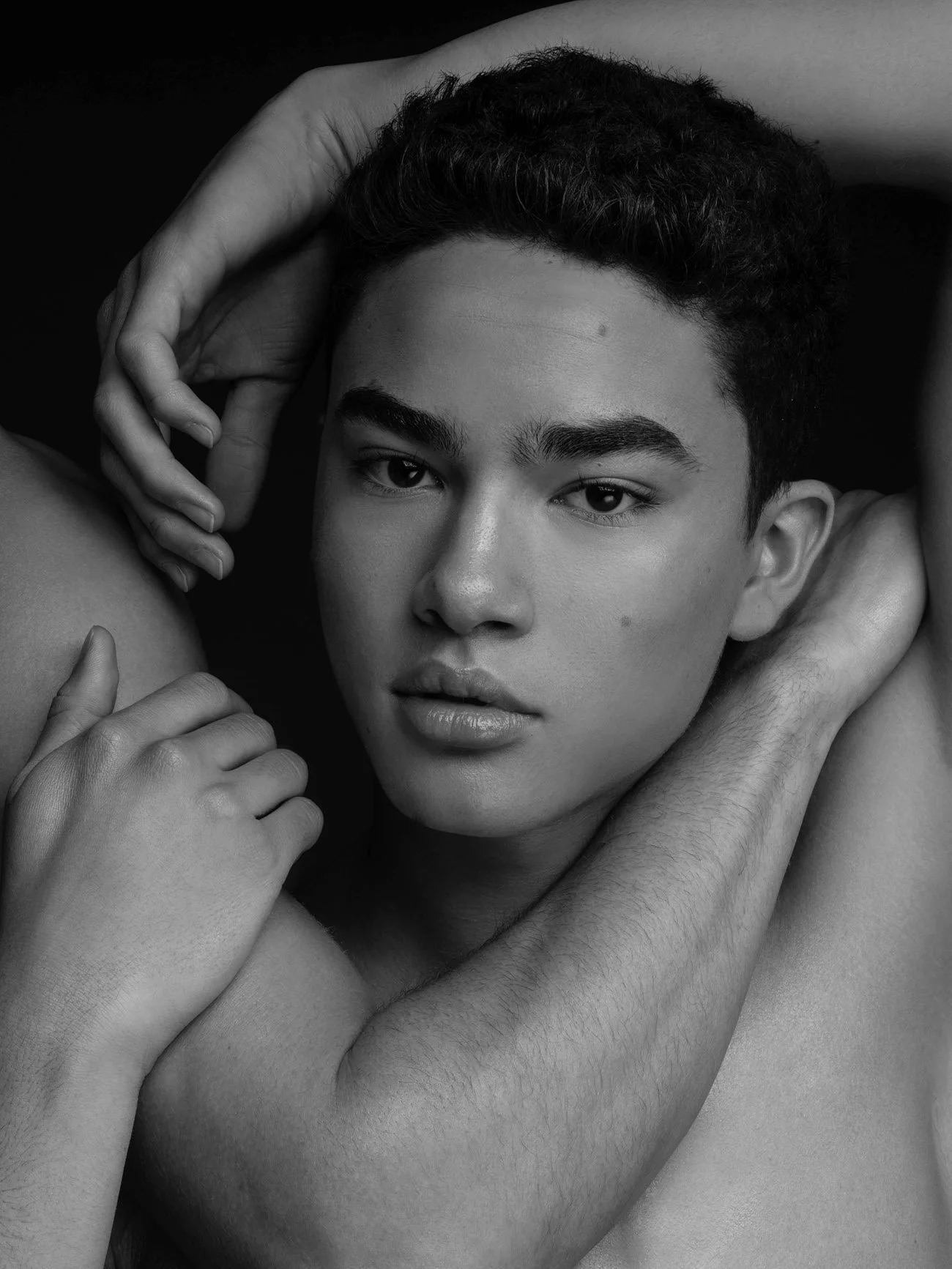 A black and white photo of a young man with short curly hair, looking directly at the camera with a neutral expression, holding his head and shoulder with his hands.