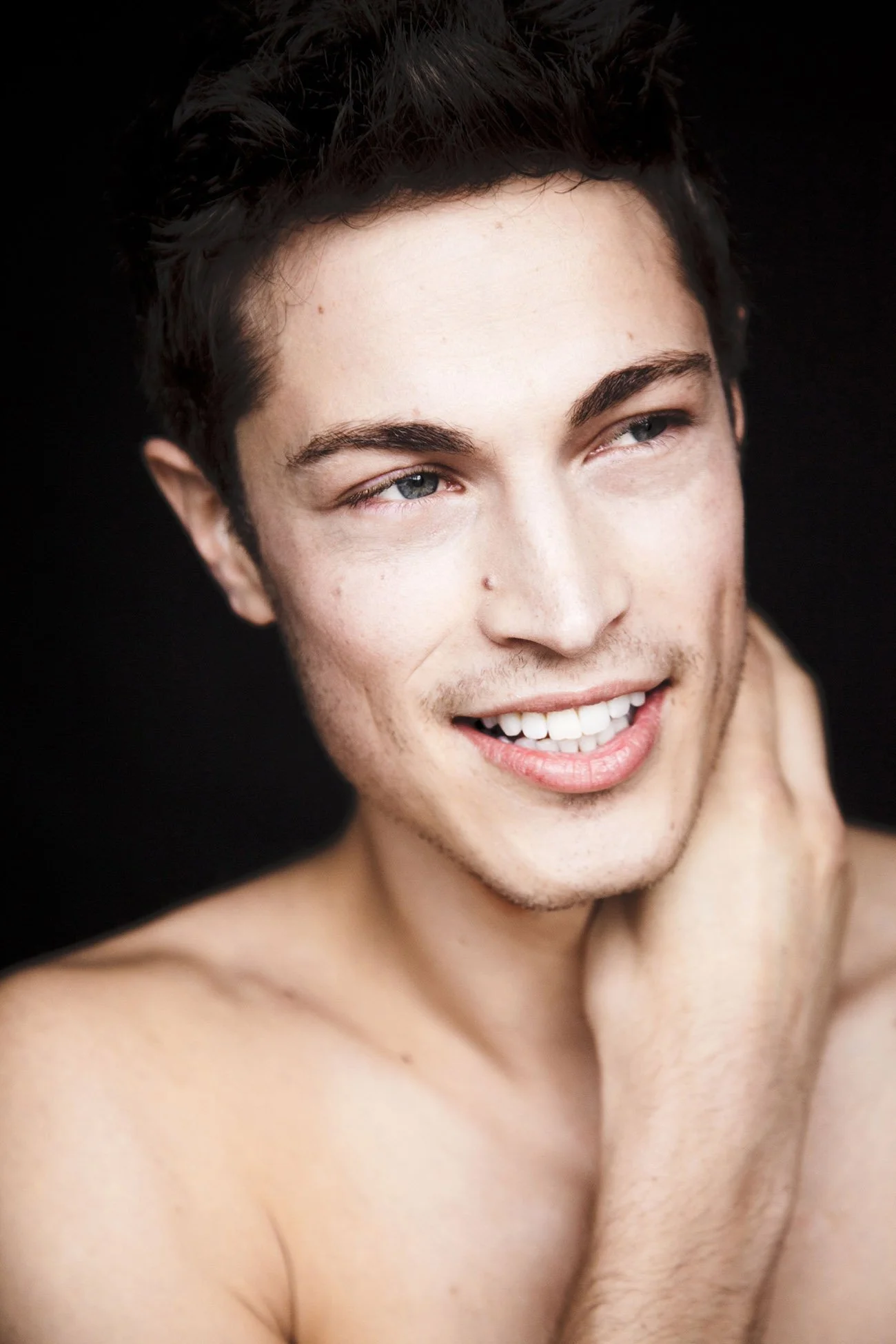 Close-up of a young man with short dark hair, blue eyes, and a light complexion, smiling and looking off to the side, touching his neck, against a black background.