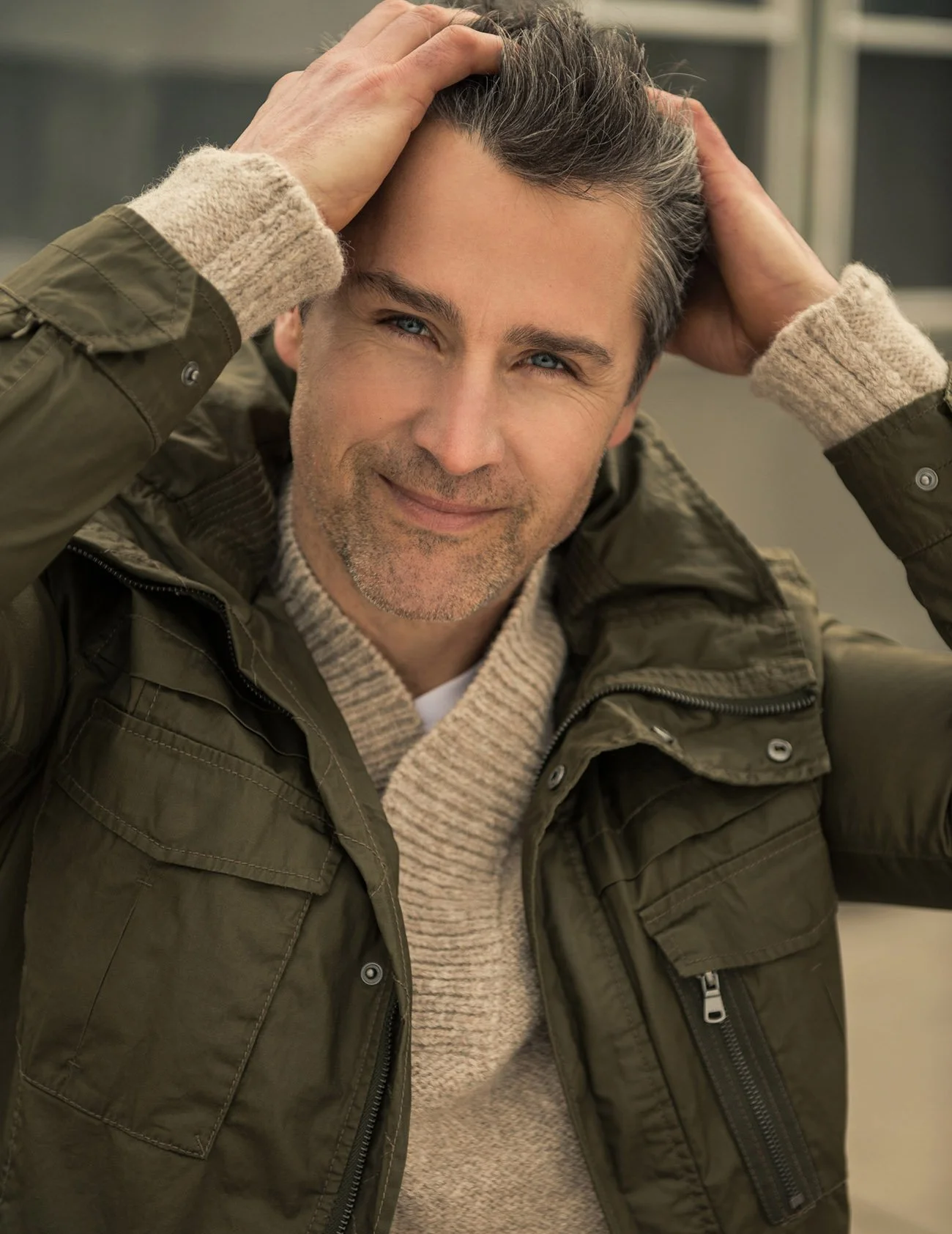 A man with gray hair and blue eyes smiling, wearing a beige knit sweater and an olive green jacket, with hands on his head.